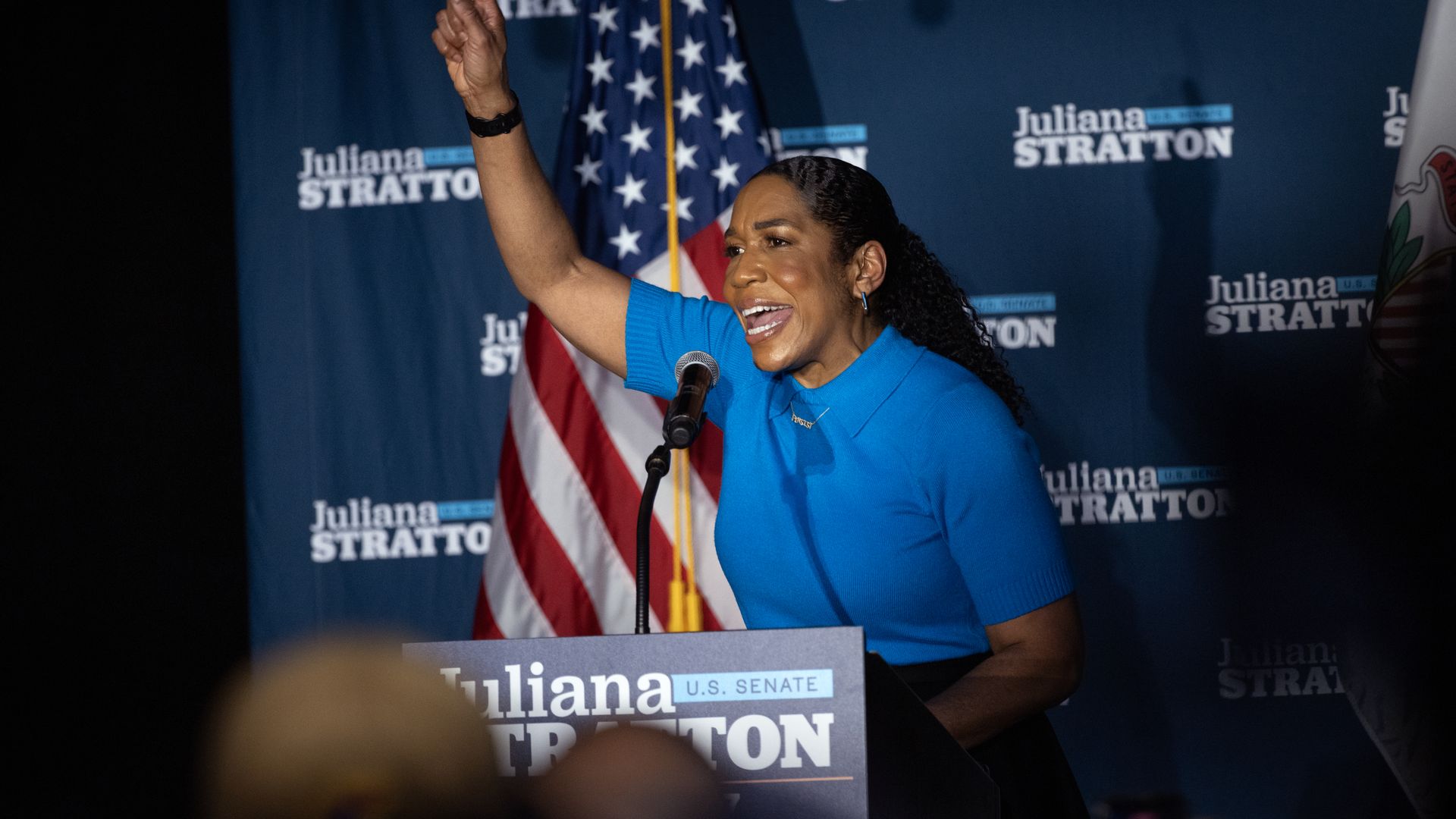 Smiling woman wearing a bright blue top raises her right fist while speaking into a microphone at a campaign rally; American flag and Juliana Stratton banners in the background.