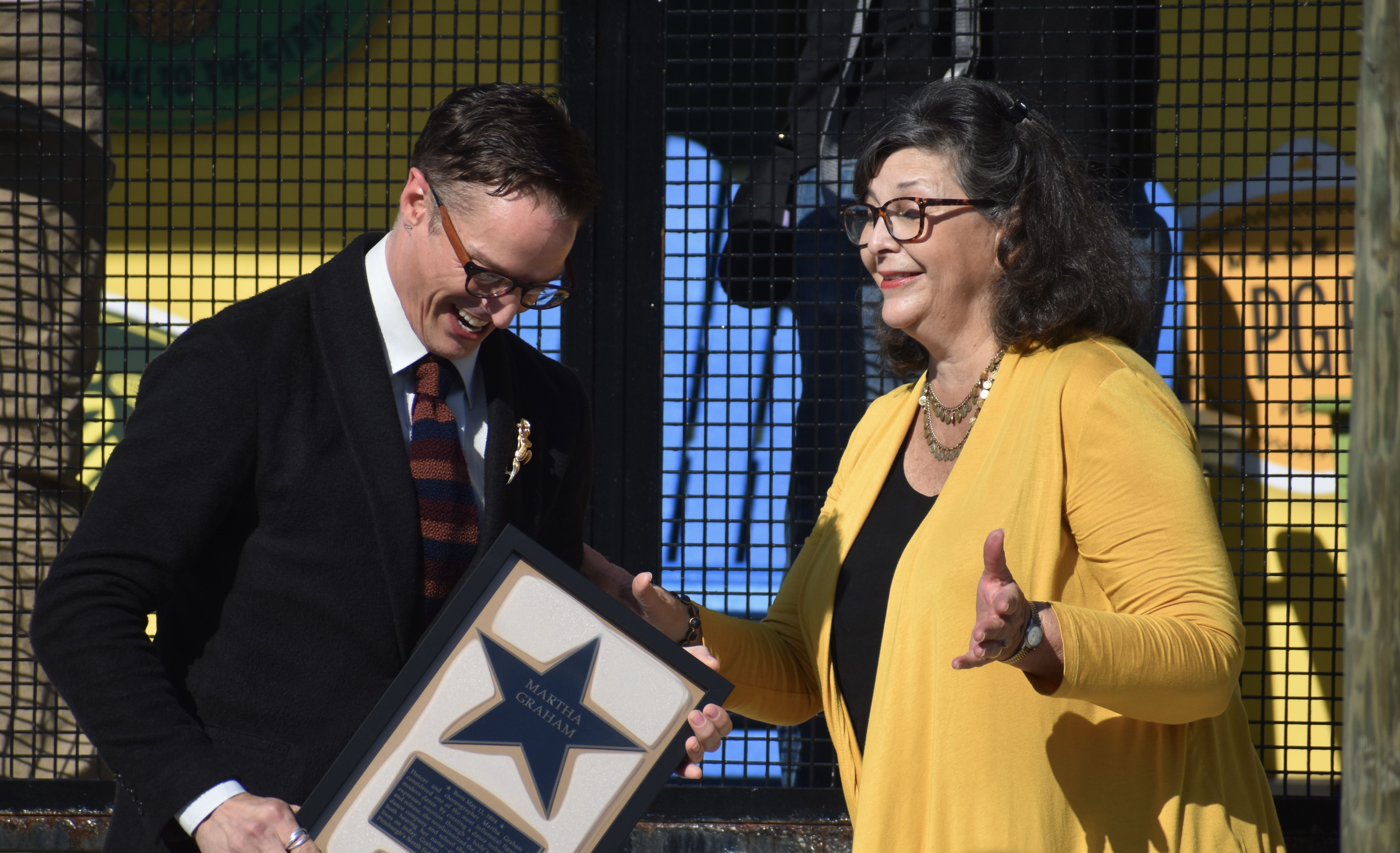 Two people outside behind a black wire fence: a man in a dark suit with a striped tie presents a framed award featuring a blue star, while a smiling woman wearing a yellow cardigan speaks.