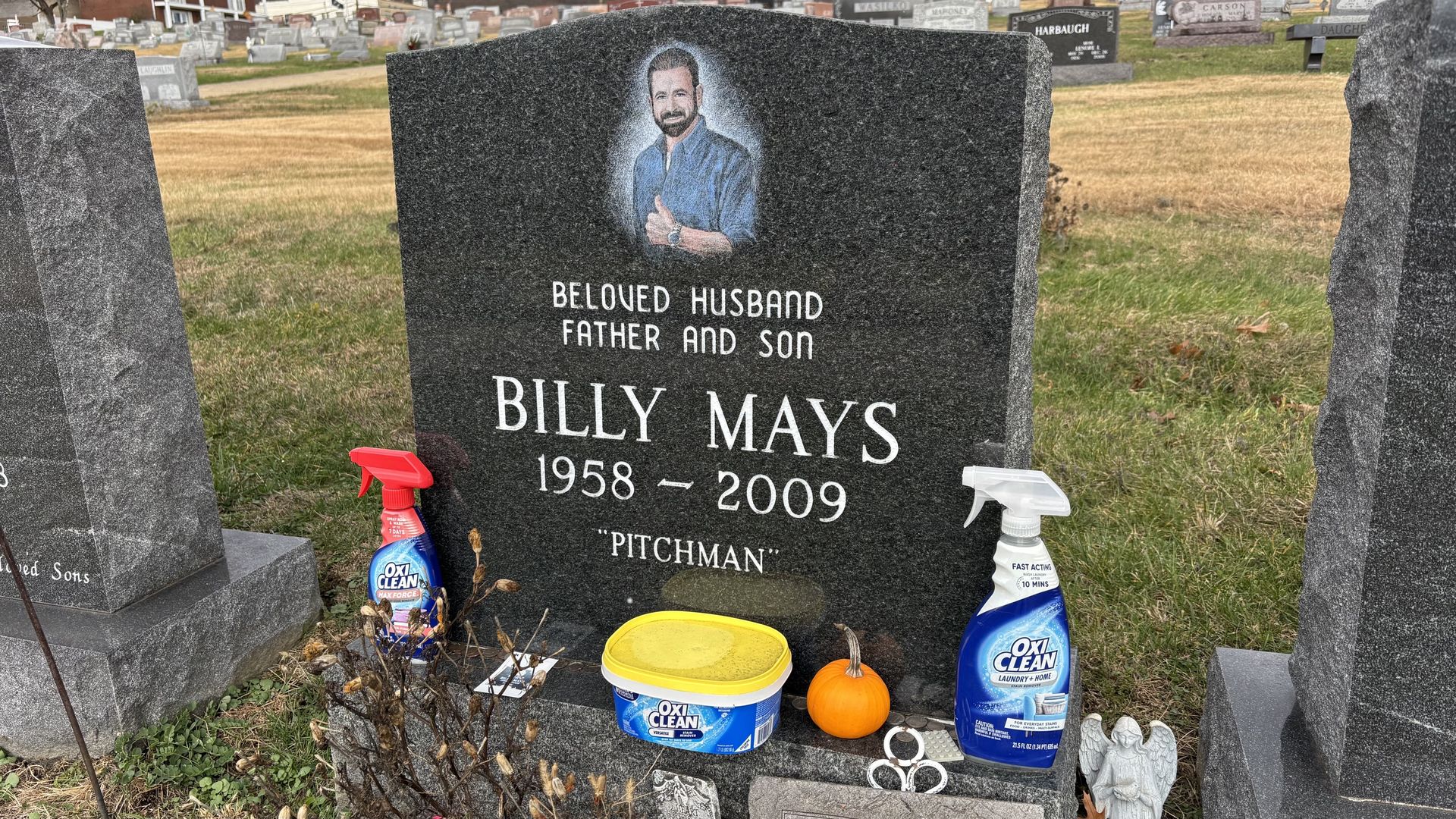 Gravestone for Billy Mays (1958-2009) with a colored portrait showing him giving a thumbs-up. Items placed in front include OxiClean products, a small pumpkin, and a small angel statue.