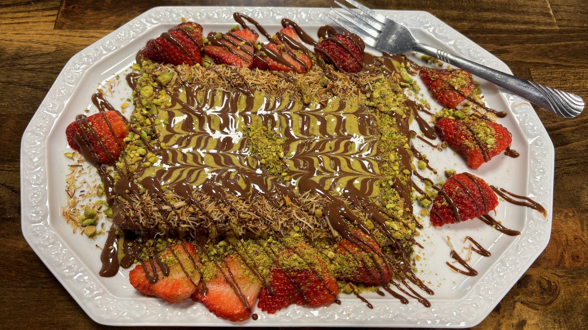 Rectangle dessert with chocolate and green pistachio drizzle, topped with shredded nuts, surrounded by sliced strawberries drizzled with chocolate on white ornate plate with fork on wooden table.
