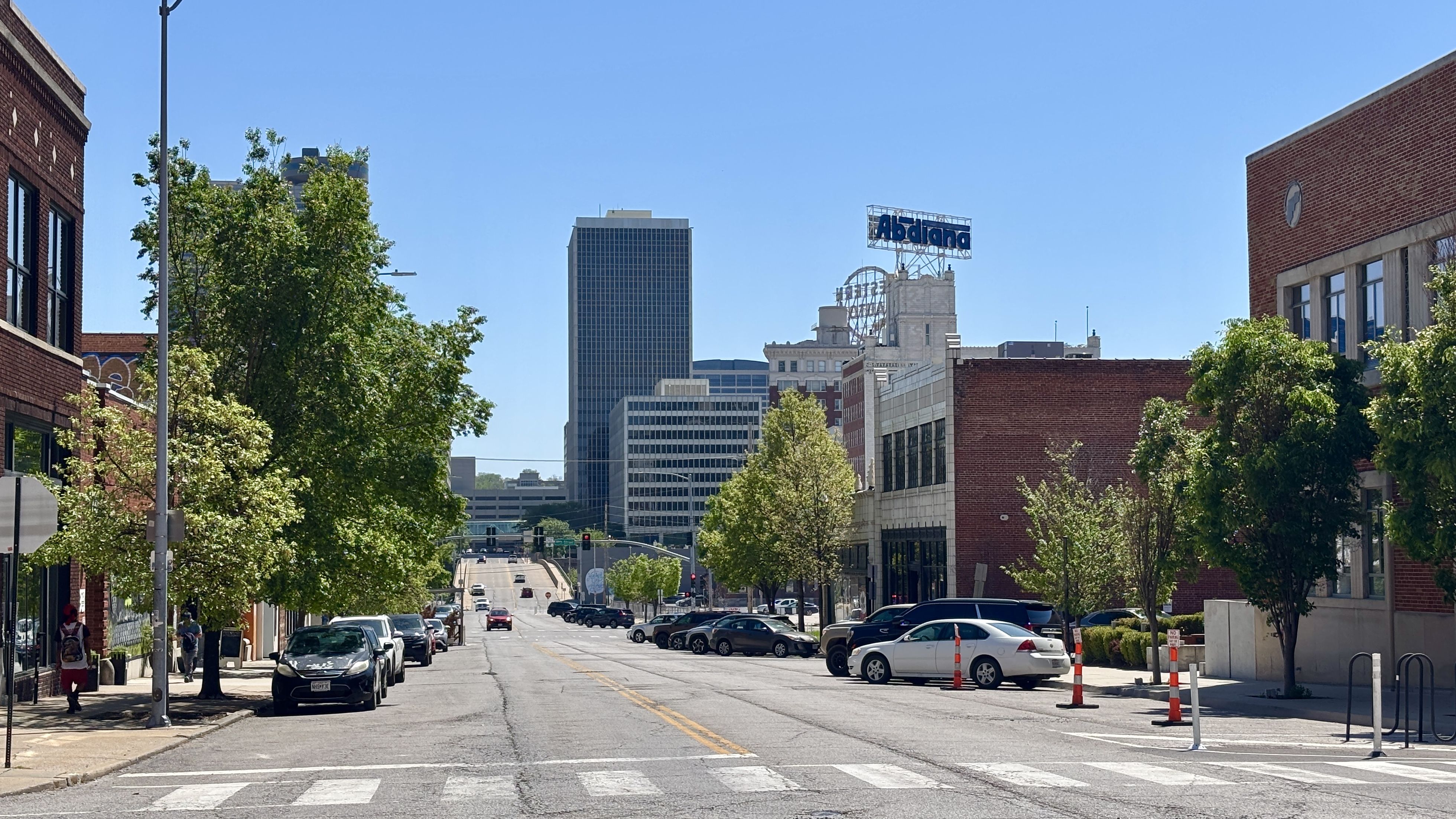Sunny city street with brick storefronts and green trees. Cars line the curb; a crosswalk and a tall glass building appear in the distance, with a blue Adriana sign atop a white building.