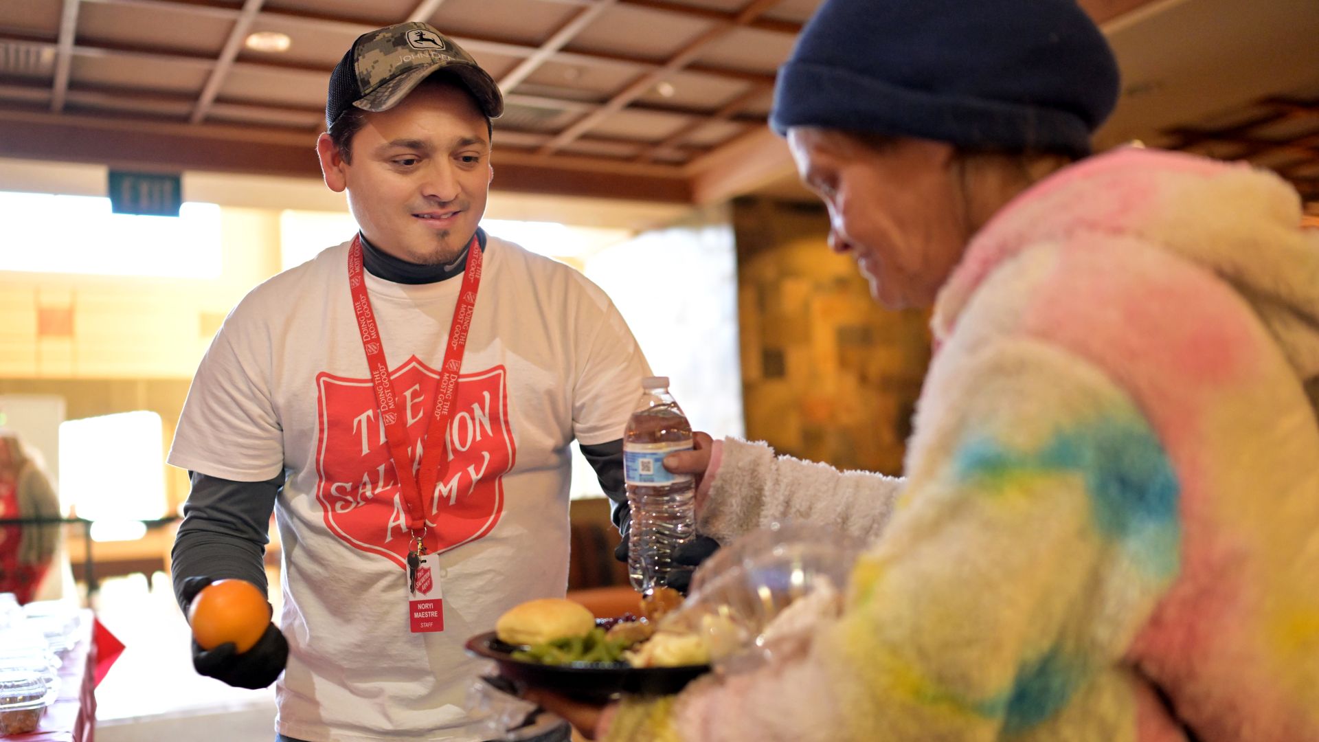 A man wearing a John Deere hat and a Salvation Army t-shirt hands an orange and water bottle to a person in a colorful fuzzy jacket holding a tray of food indoors.