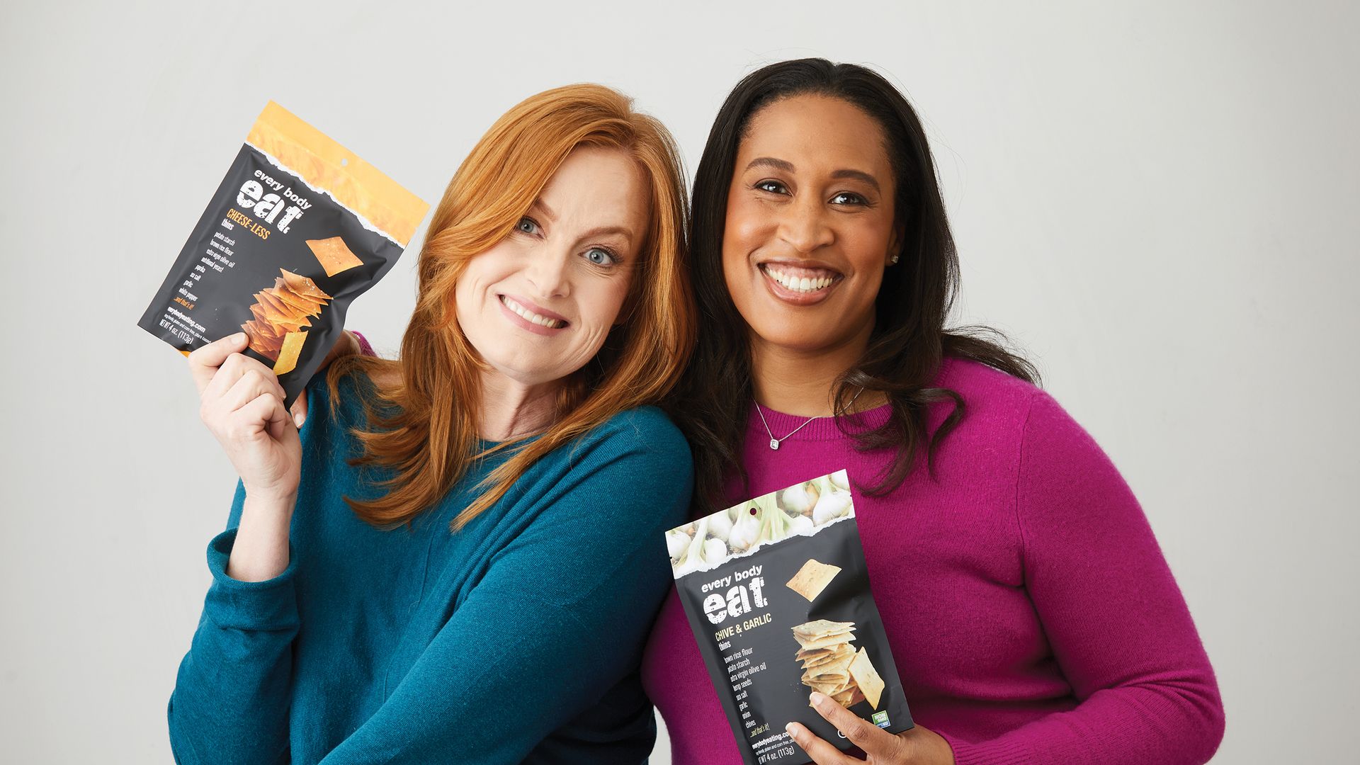 Two smiling women, one with red hair in a teal sweater, the other with dark hair in a magenta sweater, holding black and yellow snack bags labeled "every body eait" with different flavors.