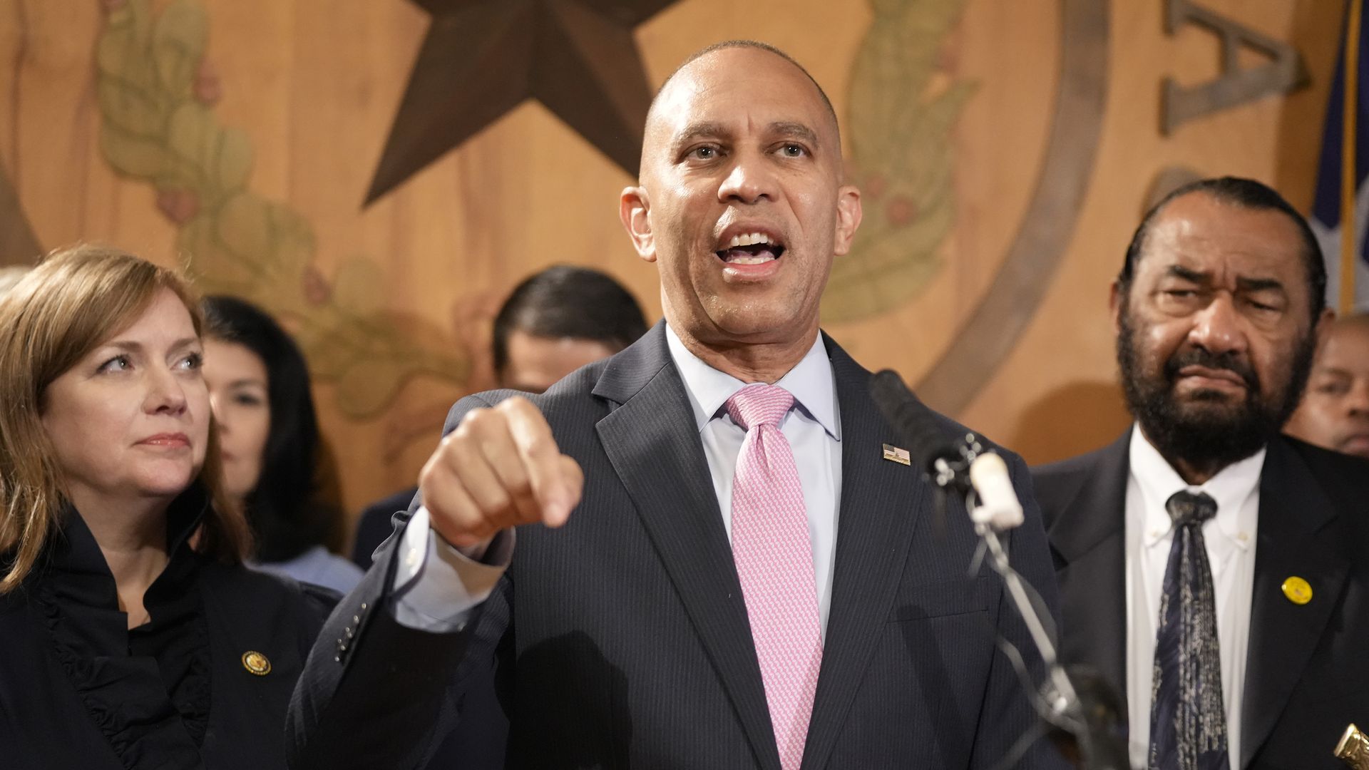 A man in a dark suit and pink tie speaks passionately at a podium with a microphone, flanked by a woman and man in suits, with a large wooden emblem featuring a star in the background.