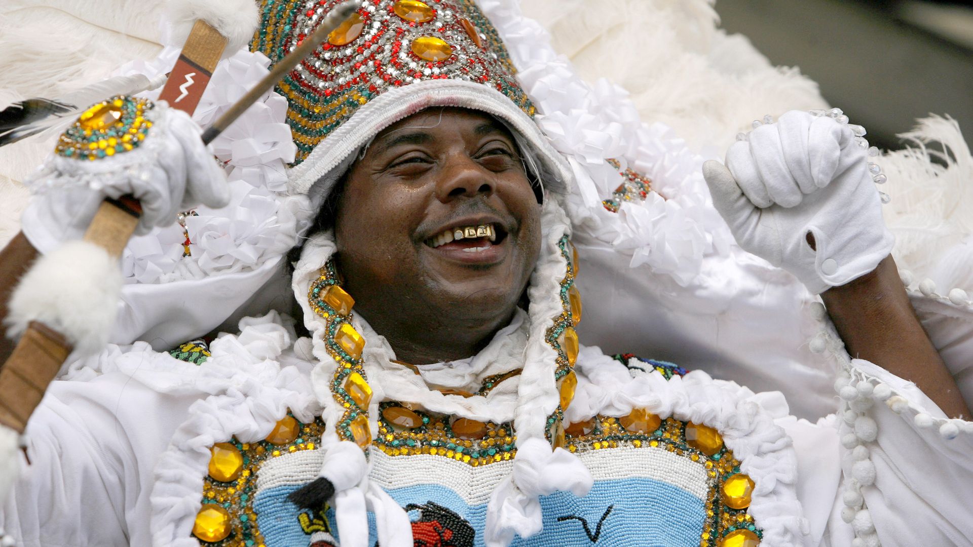 Smiling man in ornate white costume with yellow gems, beads, and feathers, wearing white gloves and gold teeth, holding a decorated stick during a festive event.