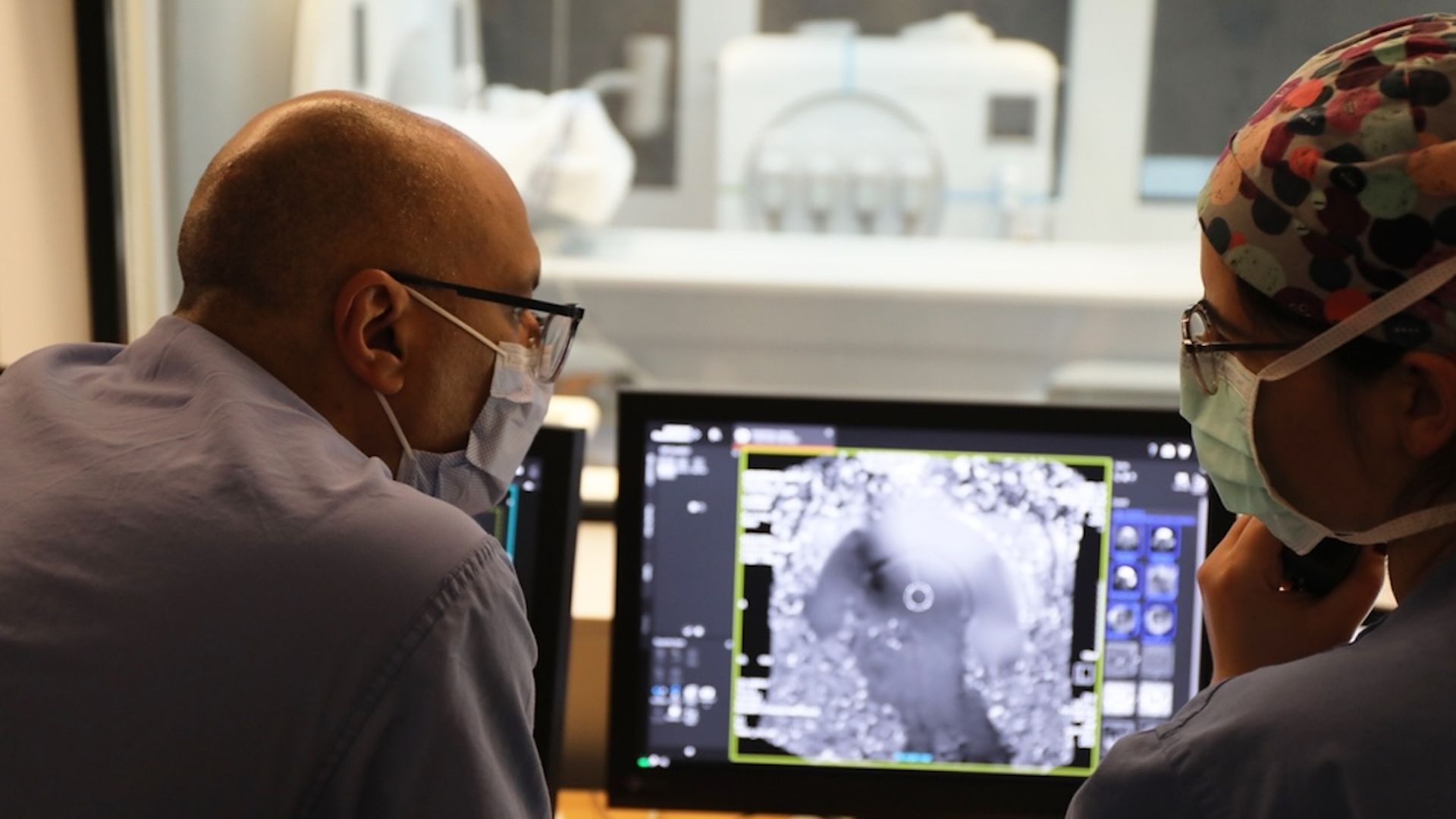 Two medical professionals wearing masks and glasses closely examine a grayscale medical scan on a computer monitor in a clinical setting.