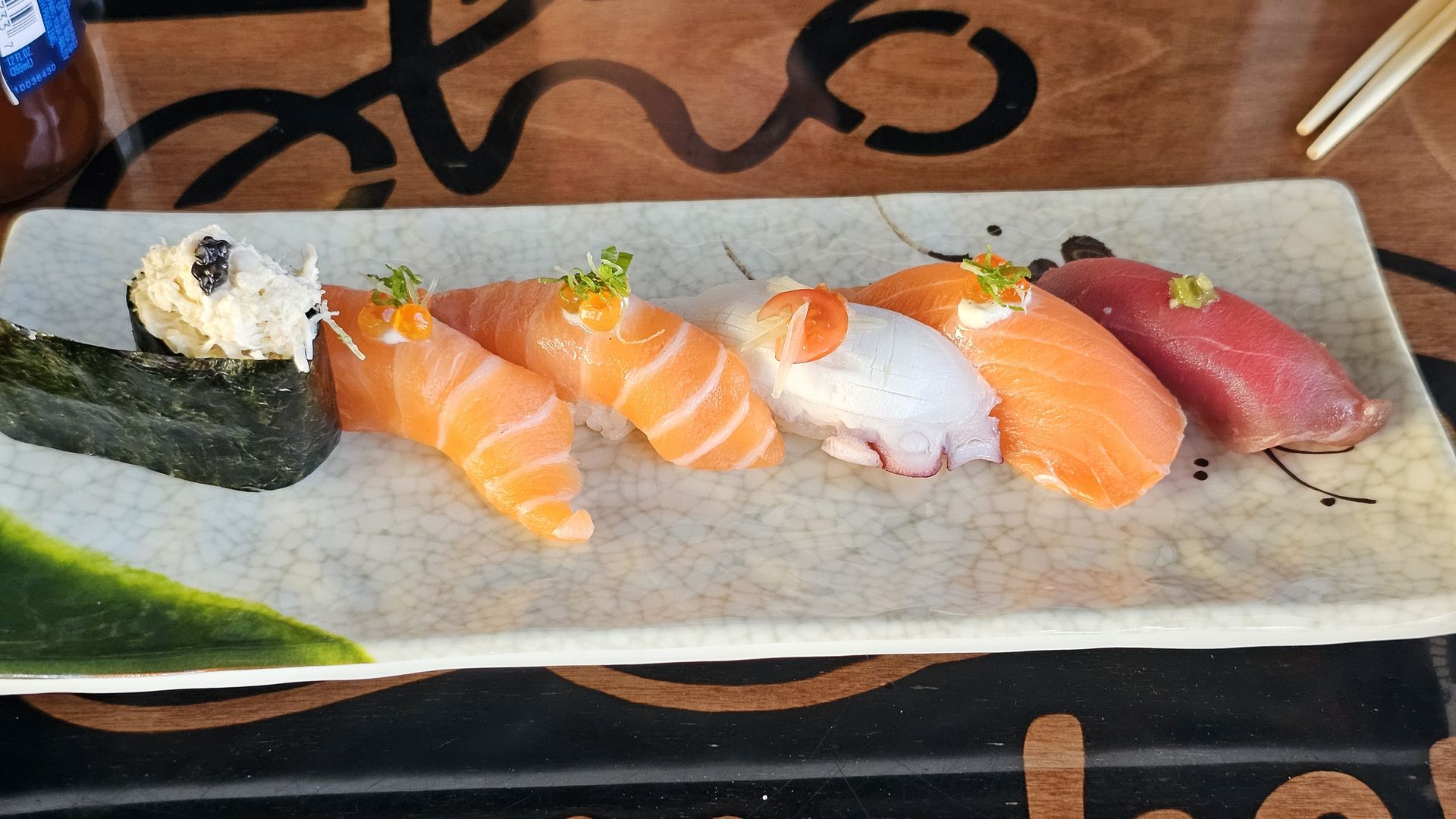 Assorted nigiri sushi on a rectangular plate: salmon, white fish, and tuna, each topped with a dab of sauce and greens; a seaweed-wrapped roll on the left, chopsticks at the top right.