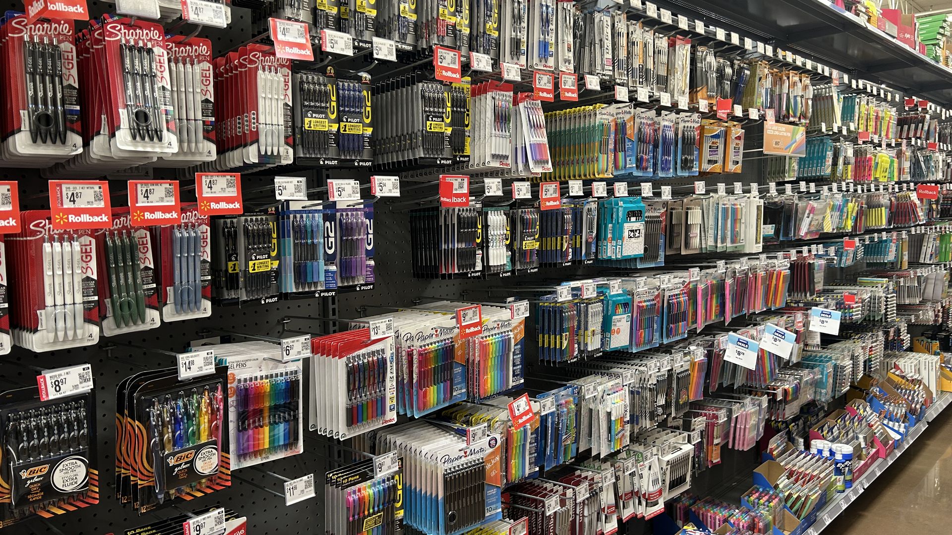 Store aisle with shelves stocked with various pens, markers, and writing instruments in colorful packaging, including Sharpie, Pilot, Paper Mate, and BIC brands, with price tags.