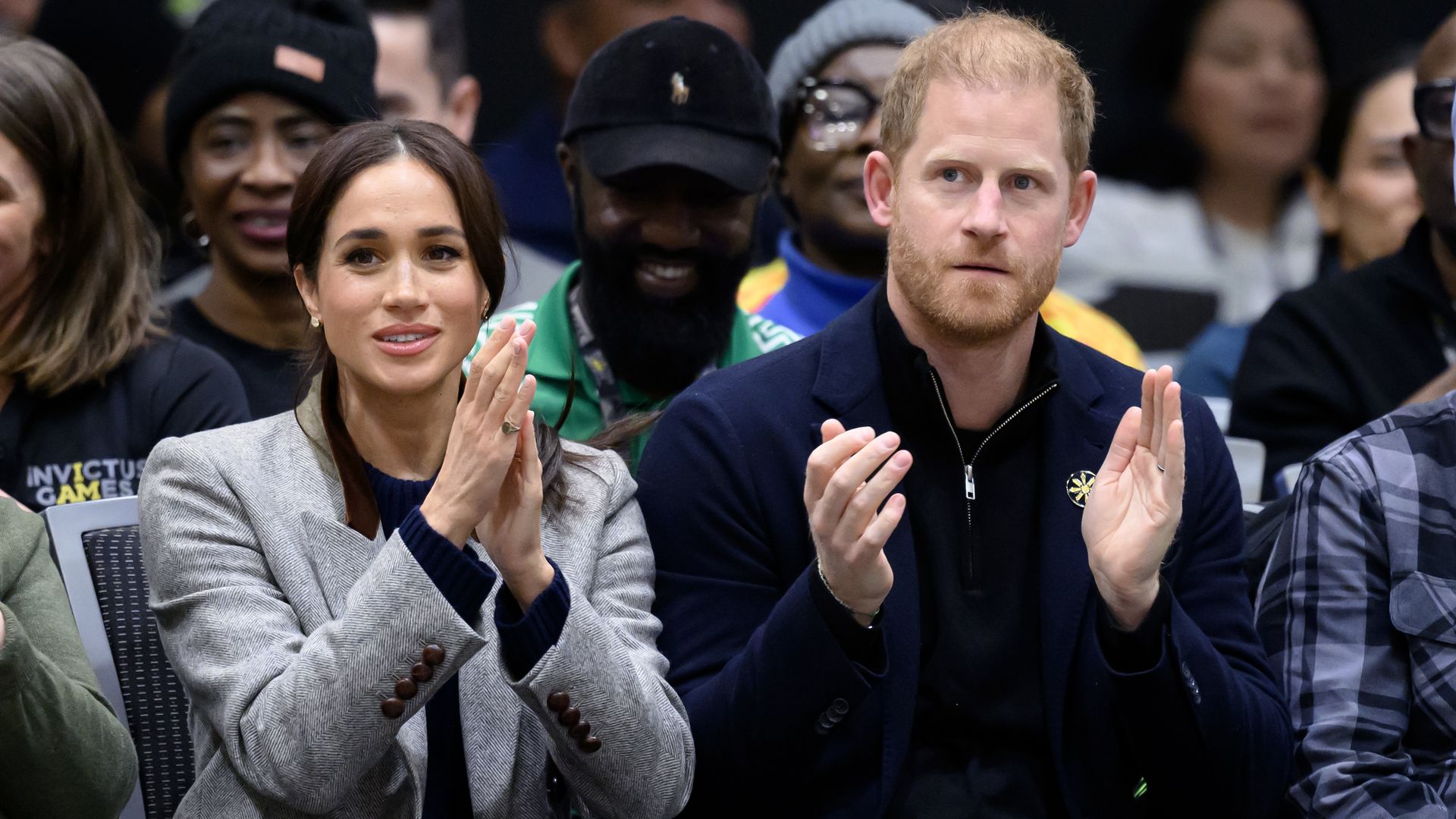A woman in a gray blazer and navy top and a man in a dark jacket clap while seated among a crowd at an event, with people wearing casual and winter attire in the background.