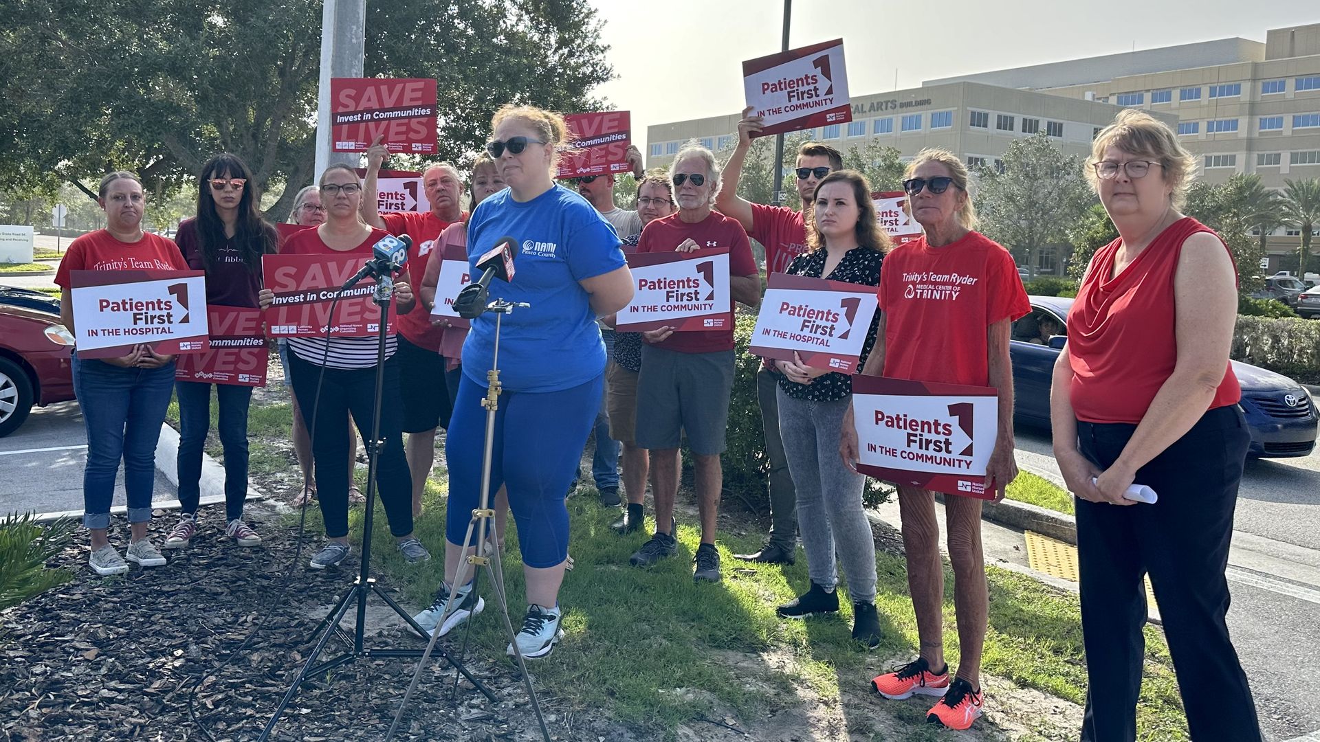 About 15 people wearing red hold "patients first" and "save lives invest in communities" signs standing behind a woman speaking into two microphones. The hospital can be seen in the background