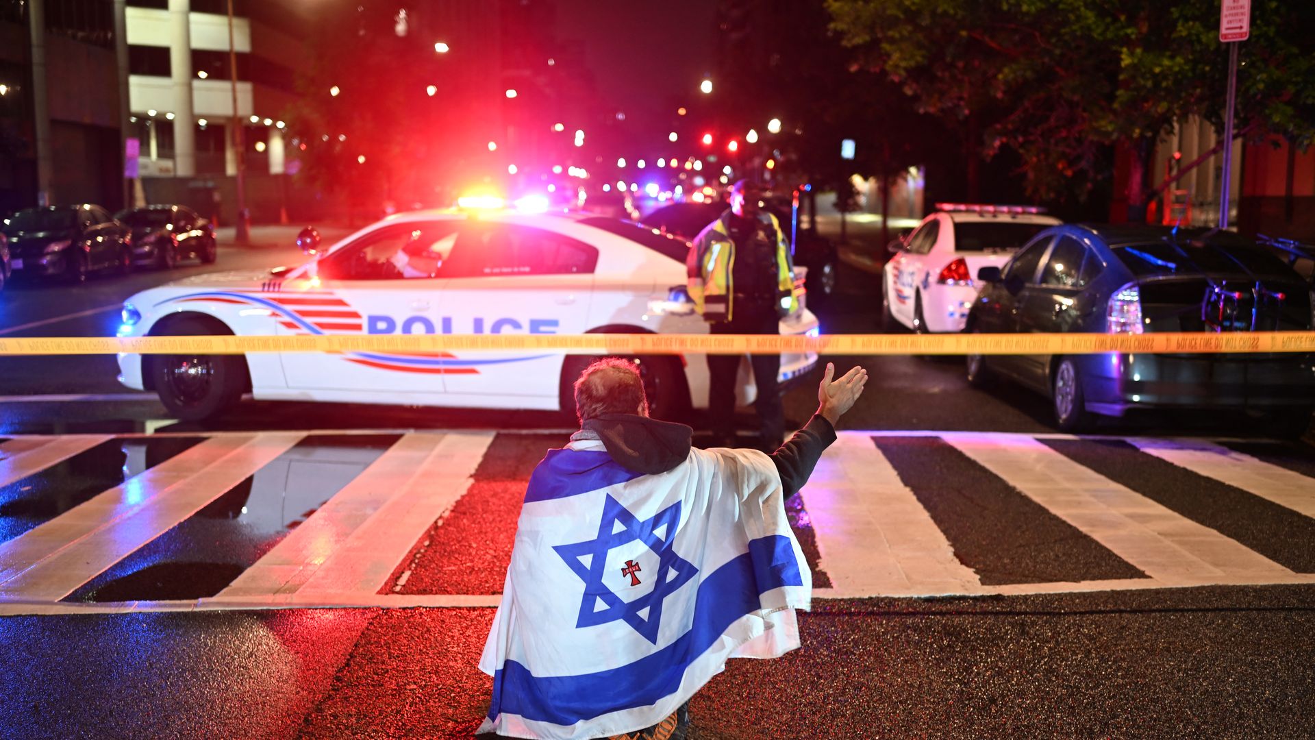 A man draped in the Israeli flag, bearing a cross and the name "Jesus" at its center, gestures as Metropolitan Police officers secure the area outside the Capital Jewish Museum following a shooting that left two people dead in Washington, DC, in the early hours of May 22, 2025. 
