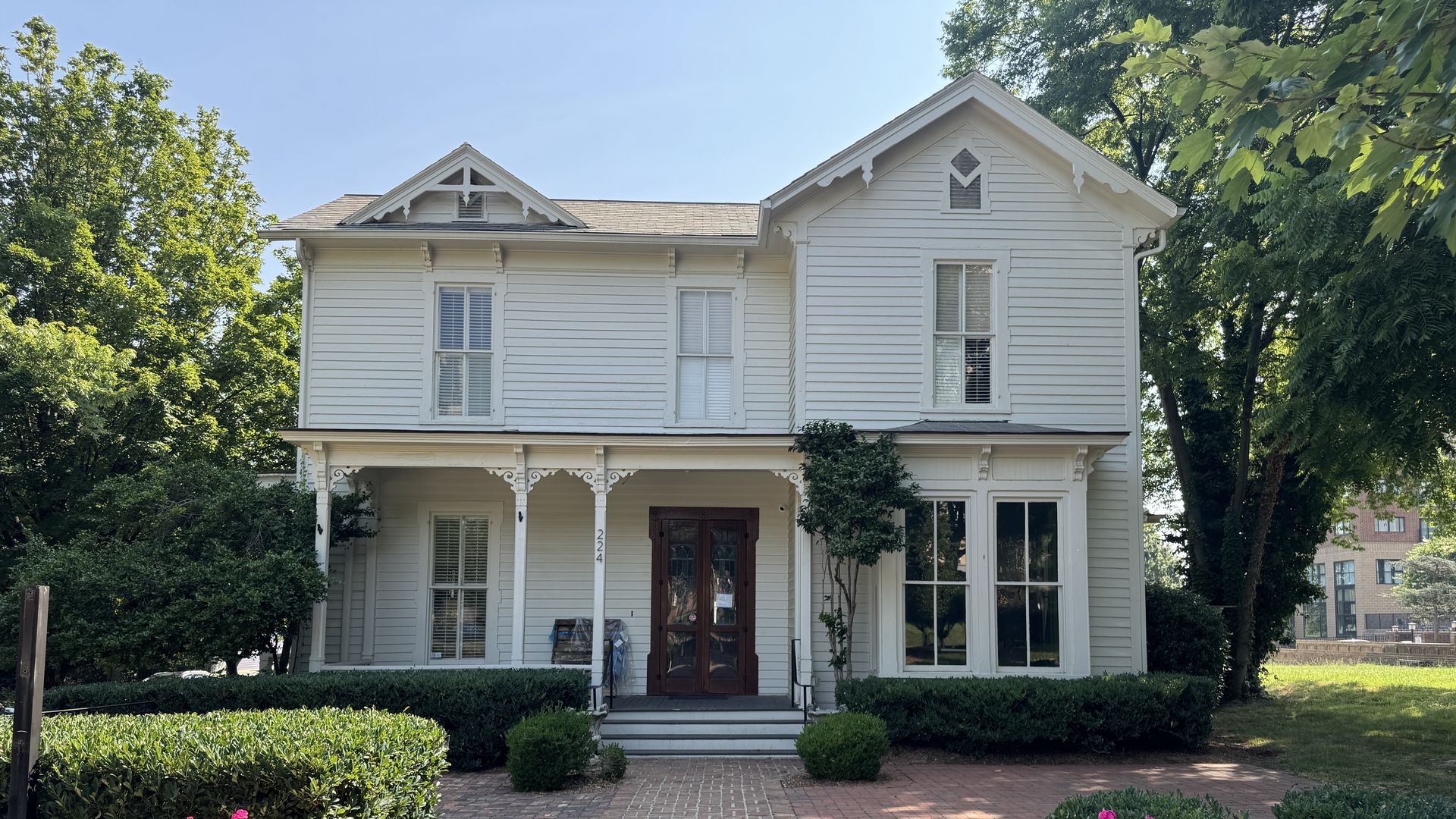 A white two-story house with a pitched roof and decorative wooden trim around the top. The facade has large windows and a central door, flanked by neatly trimmed bushes. A brick pathway leads to the entrance, and two large planters with vibrant pink flowers are positioned symmetrically on either sid