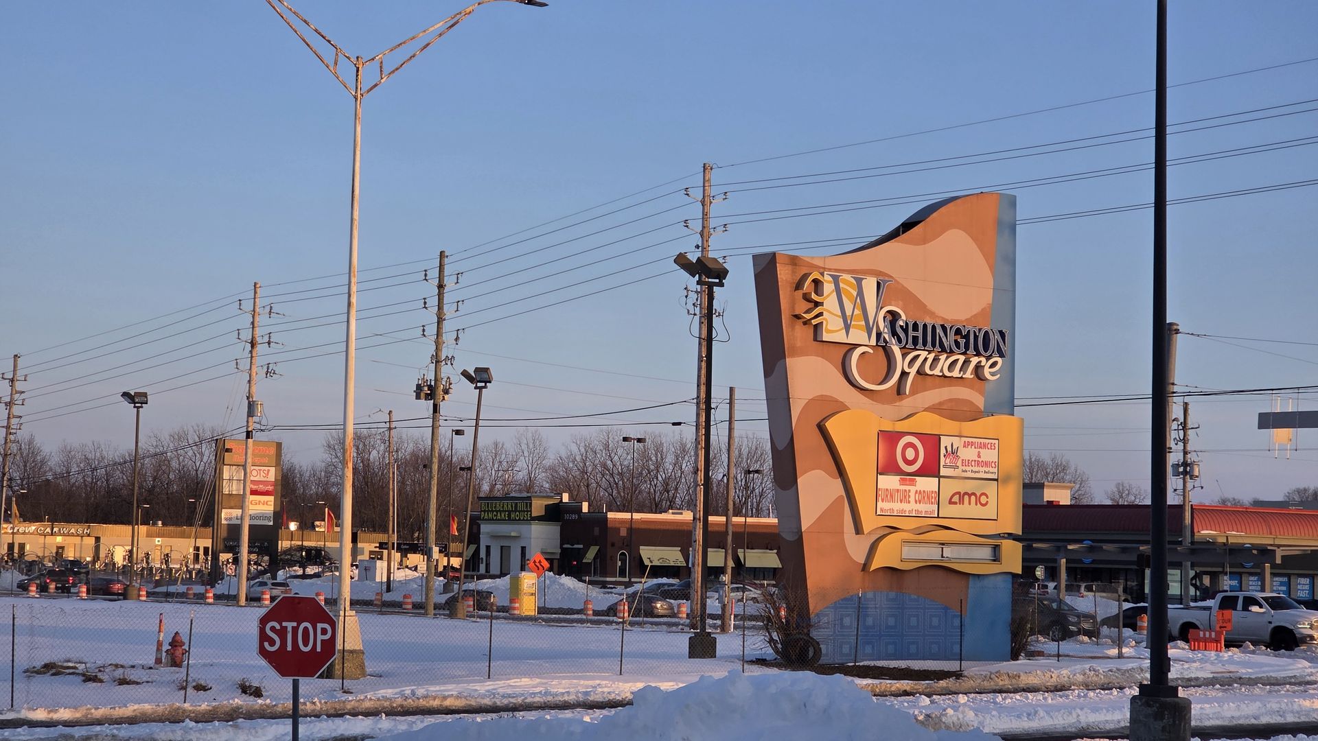 Wide view of a shopping plaza with a tall curved sign reading "Washington Square," many utility poles and power lines, a clear blue sky, snowy ground, and a stop sign in the foreground.