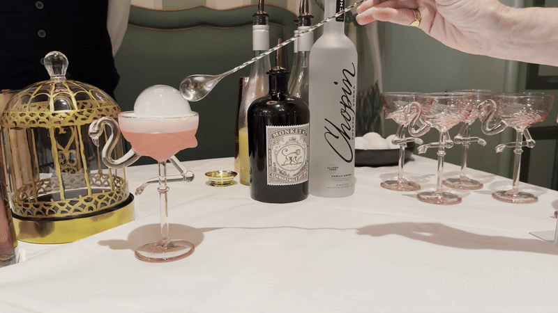 A person pops a bubble with a spoon as it sits on top of a cocktail in a flamingo-shaped glass.