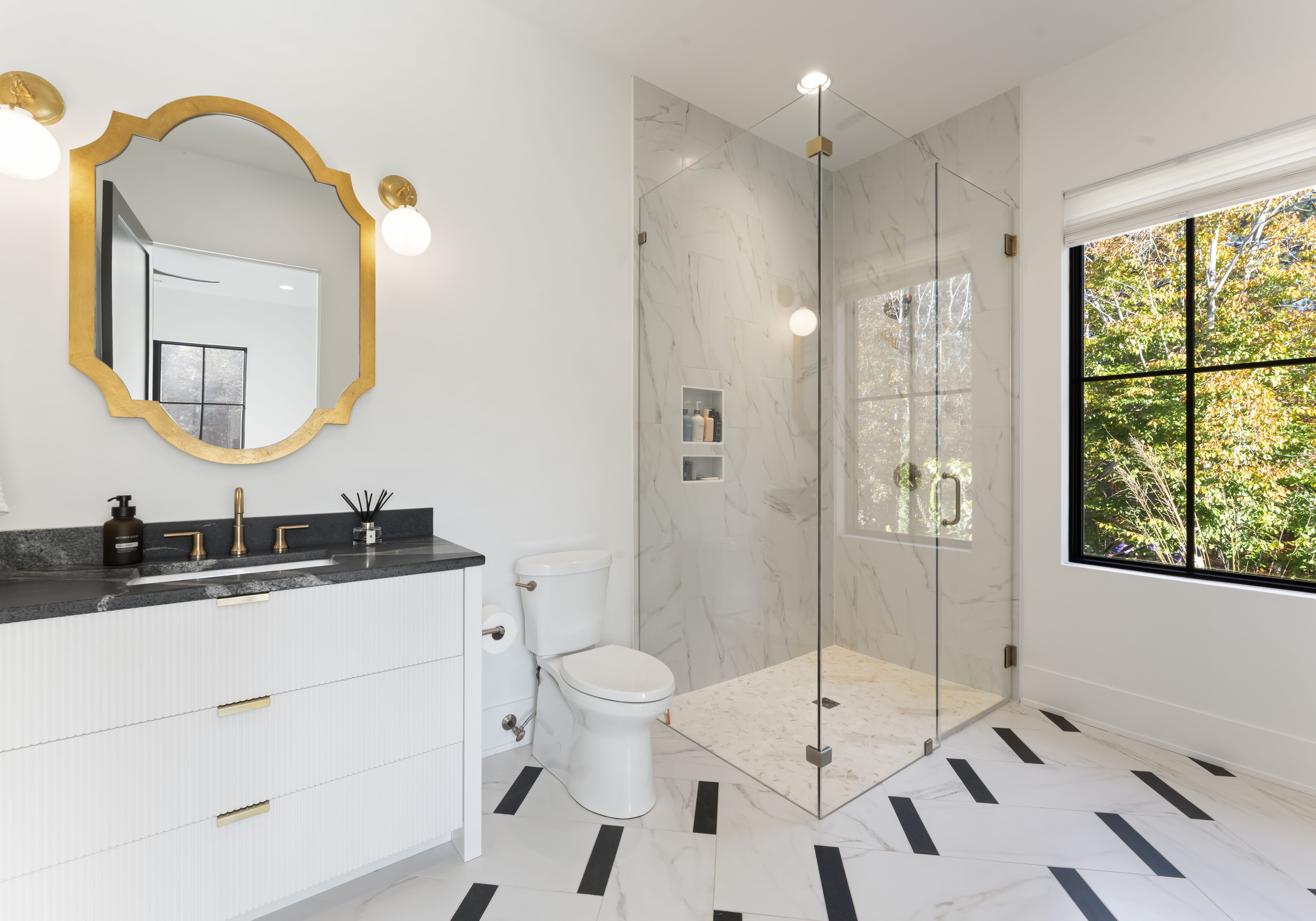 Bright white bathroom with a gold scalloped mirror, globe sconces, black countertop, white vanity, brass faucet, toilet, and a glass shower with marble tiles; a large window shows green trees outside.