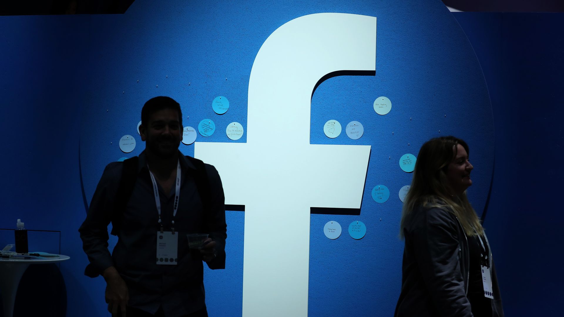 In this image, a man and a woman wearing event lanyards stand on either side of a large Facebook "F" logo.