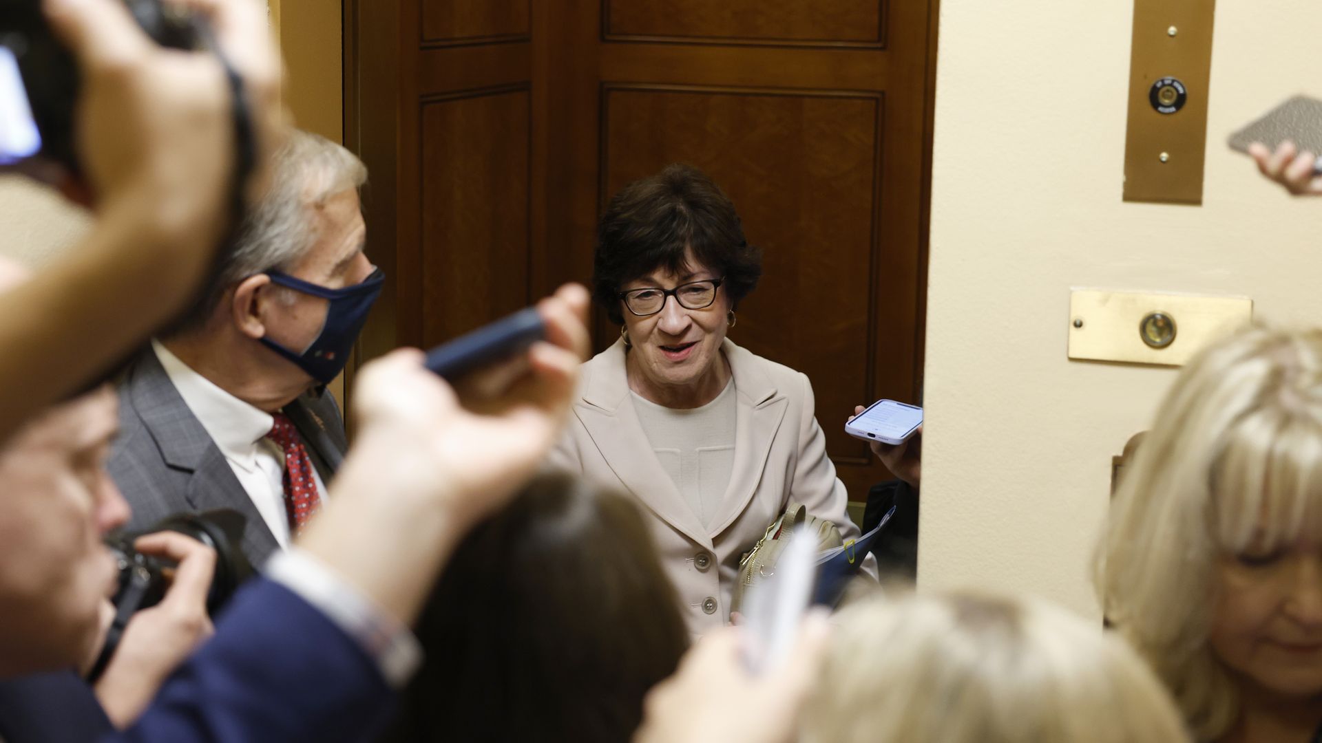 Sen. Susan Collins, wearing a white shirt and white jacket, stands in a Senate elevator while talking to a throng of reporters. 
