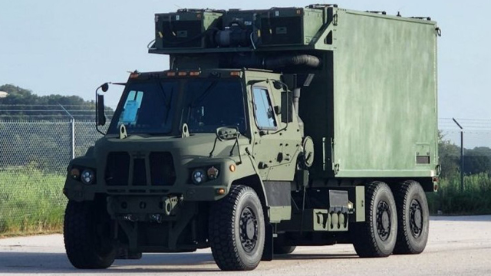 A large green military vehicle sits on a road at Joint Base Lewis-McChord. The sky is blue. Grass can be seen in the back. A fence lines the area.