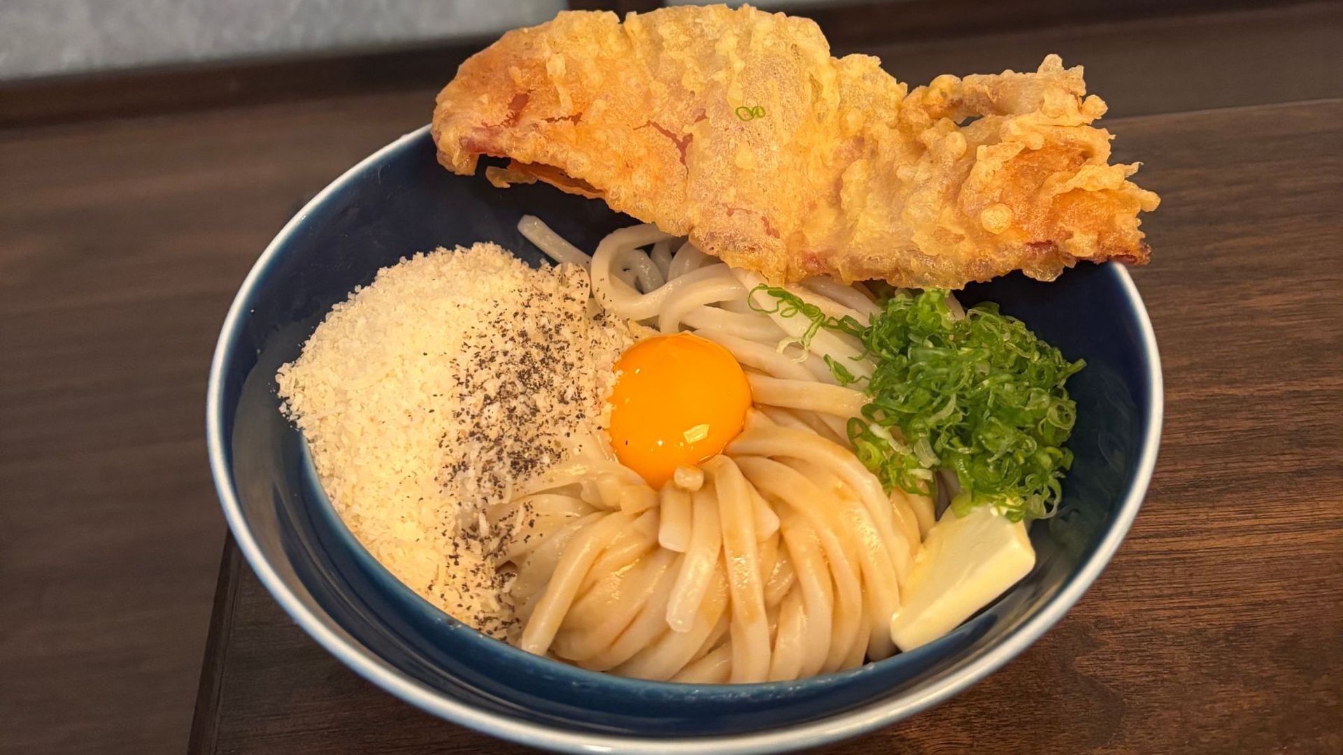 Blue bowl of udon noodles topped with a raw egg yolk, green onions, and a large crispy tempura on a dark wooden table.