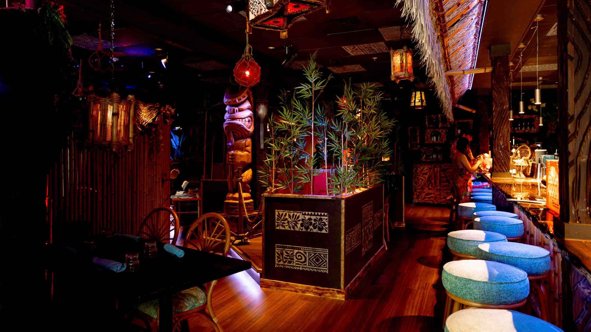 Dim tropical bar interior with bamboo, thatch, a tiki statue, hanging lanterns, and potted plants; a row of blue-and-white cushioned stools along a wooden counter under warm red lighting.