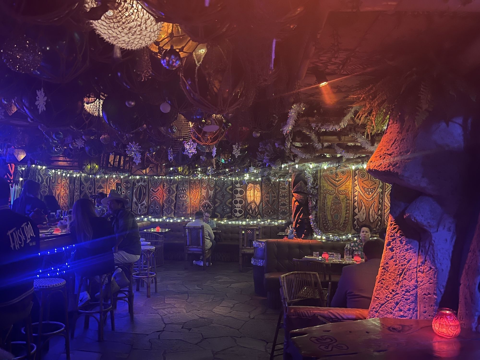 Dimly lit tiki bar with tribal wall art, wooden stools, and string lights. Patrons sit at the bar and booths under a ceiling decorated with hanging ornaments and snowflakes, bathed in blue and red light.
