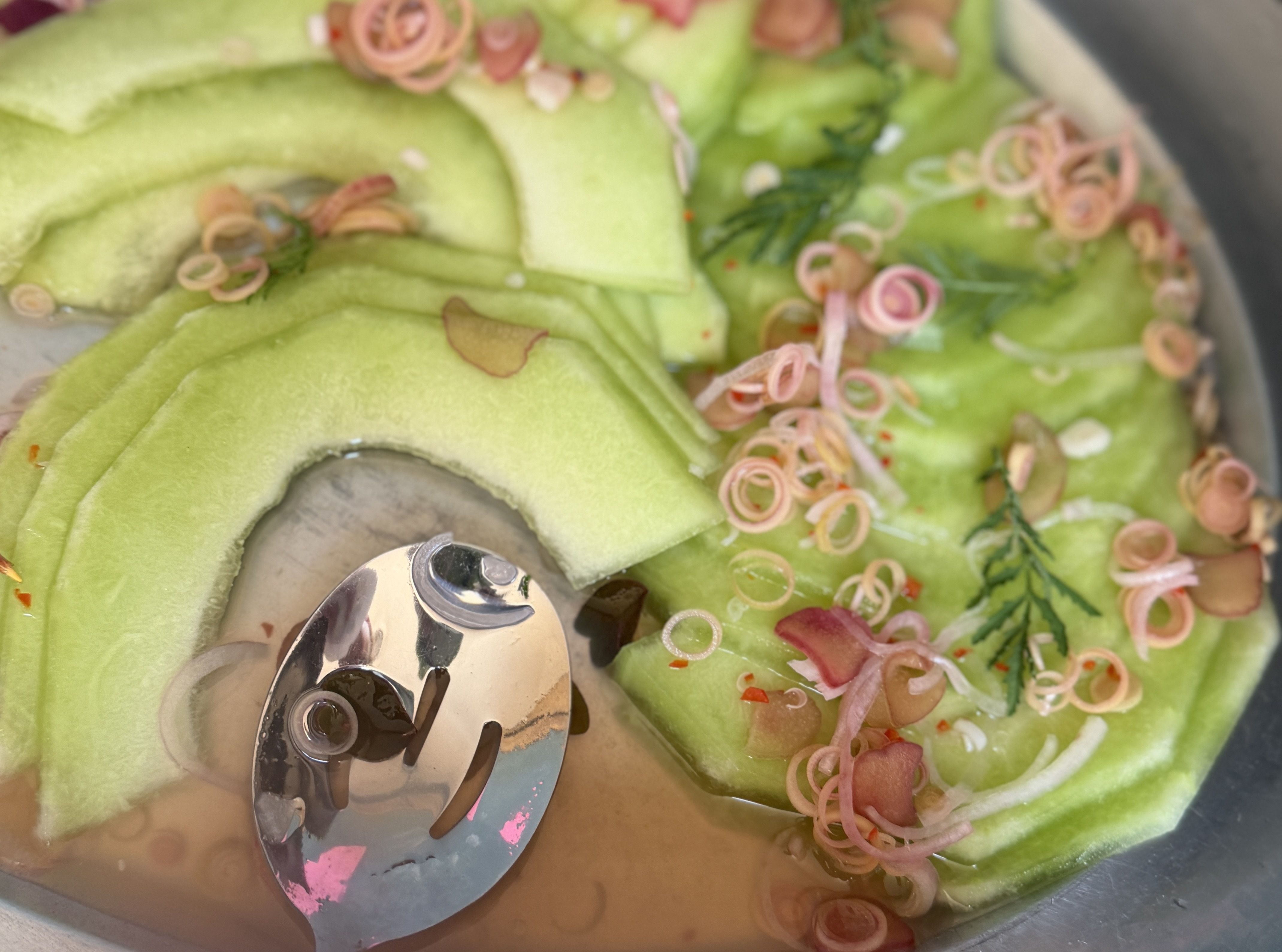 Close-up of a bright green dessert or melon slices topped with pink spiral garnishes and fresh herbs, with a shiny metal serving utensil in a bowl.