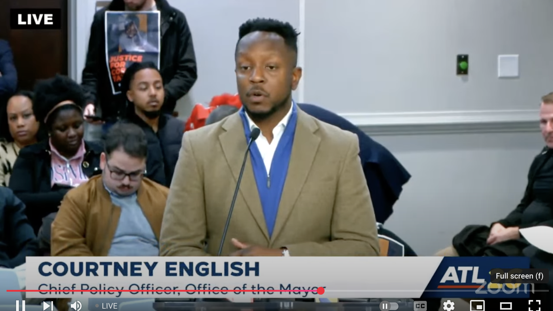 An individual wearing a tan jacket over a blue zip-up sweater speaks at a public government meeting