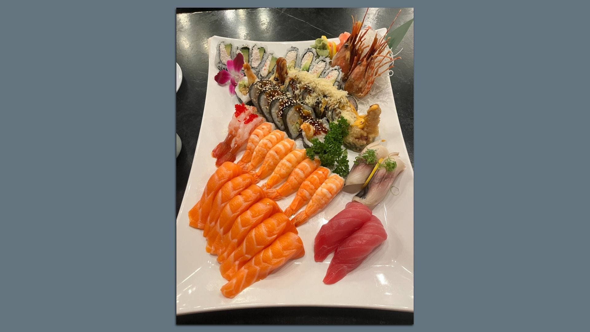 Platter of assorted sushi on a white plate: rows of salmon nigiri, tuna sashimi, shrimp, and various rolls topped with sesame; garnished with parsley and a pink orchid on the side.