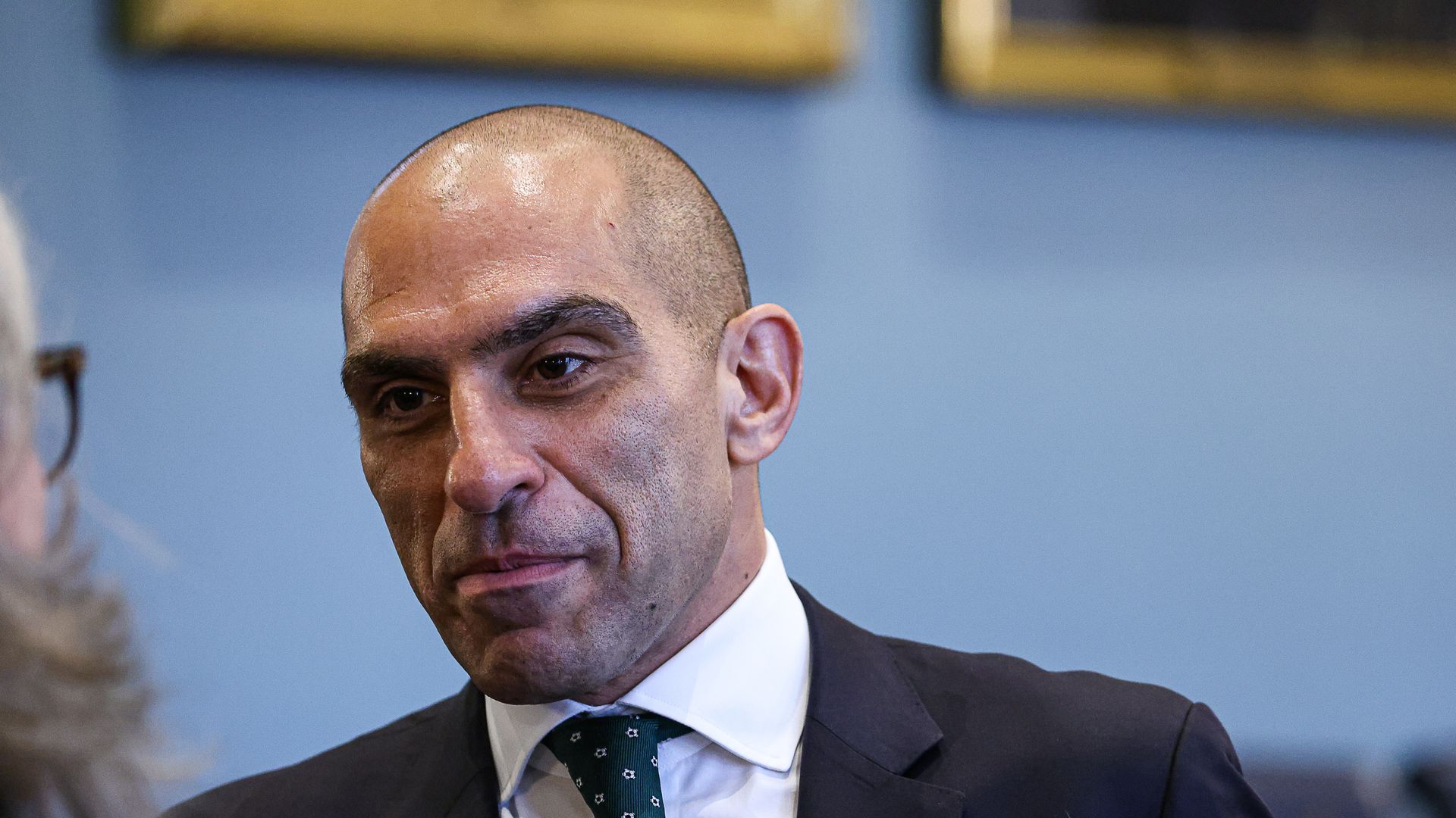 A man seen from shoulders up. Bald. In a dark suit, with a tie. Official looking room.