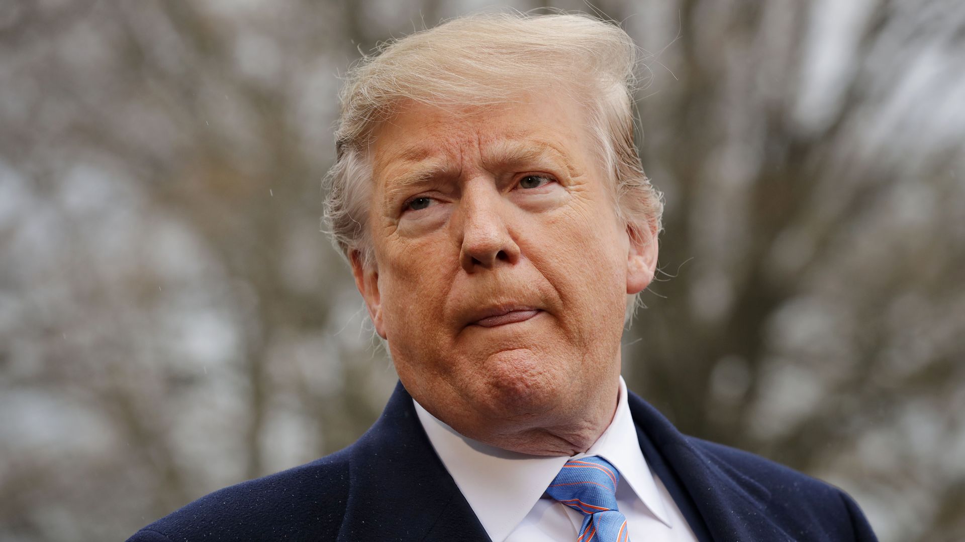 In this image, Trump stands outside and listens while wearing a suit.