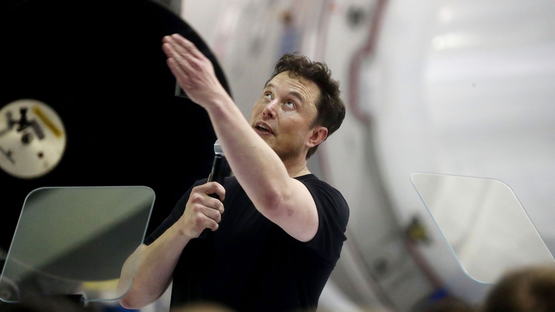 Elon Musk at a press conference pointing up at something with one hand and holding a microphone in the other.