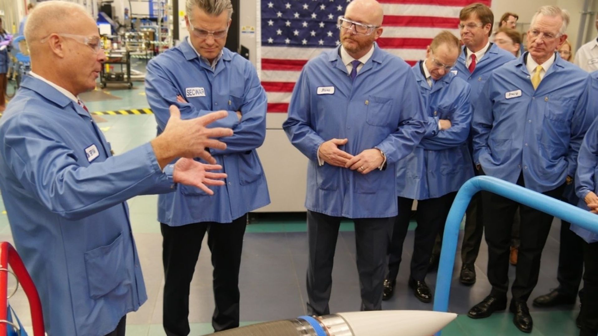 Group of men in blue lab coats and safety glasses stand around a rocket nose cone on a red platform inside a tech facility; an American flag hangs behind as one man speaks.