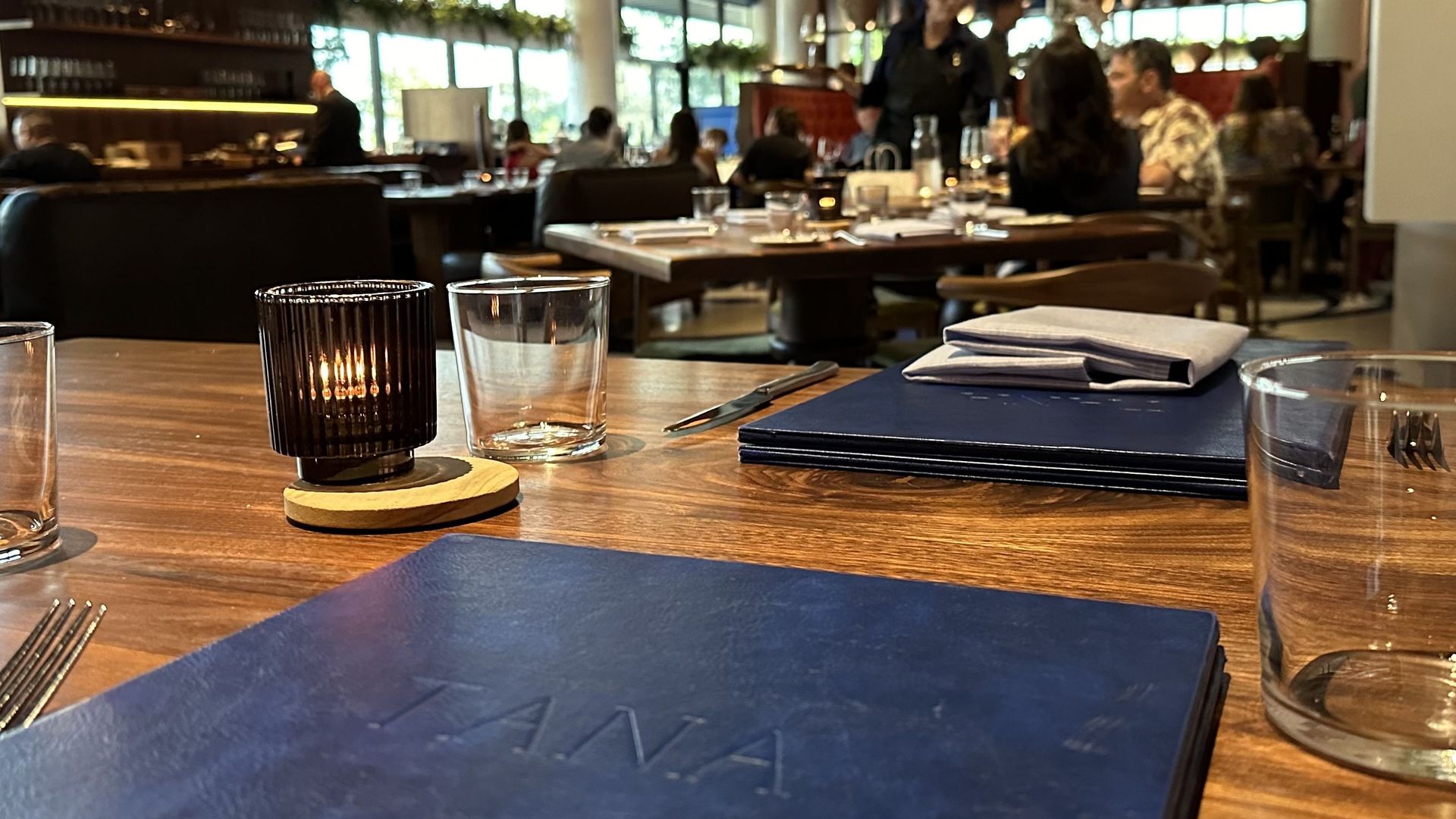 Cozy restaurant interior seen from a wooden table with a lit candle in a black ribbed glass, blue menus, and glassware; diners and staff bustle under warm pendant lights by large windows.