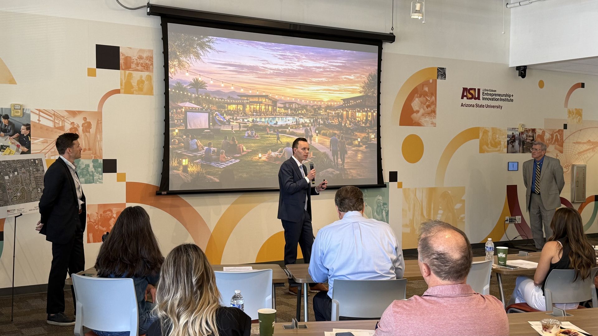 Conference room presentation with a large screen showing a sunset pool scene and string lights. A man in a suit speaks into a microphone as another man stands nearby; attendees seated, ASU branding on the walls.