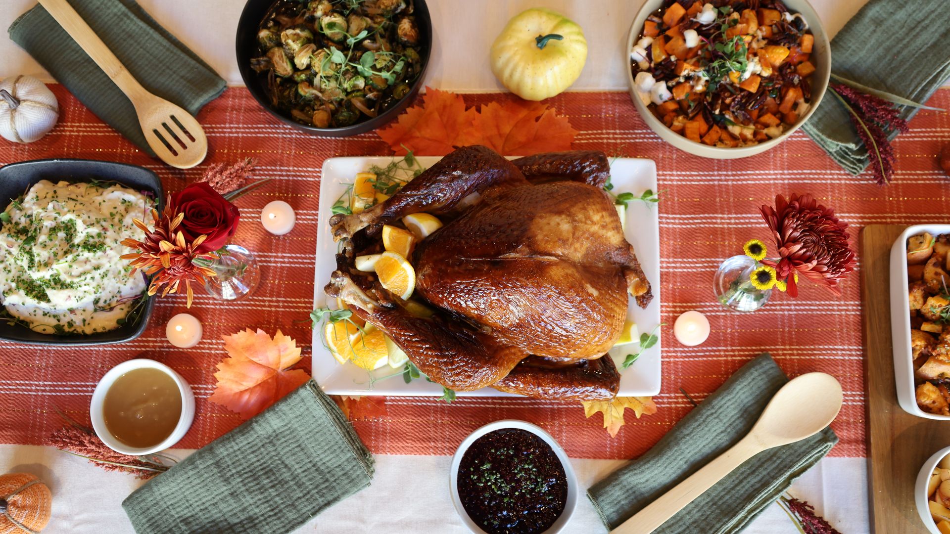 Thanksgiving table with a roasted turkey garnished with orange slices, bowls of mashed potatoes, Brussels sprouts, sweet potatoes, cranberry sauce, gravy, and green napkins on an orange plaid runner with fall leaves and candles.