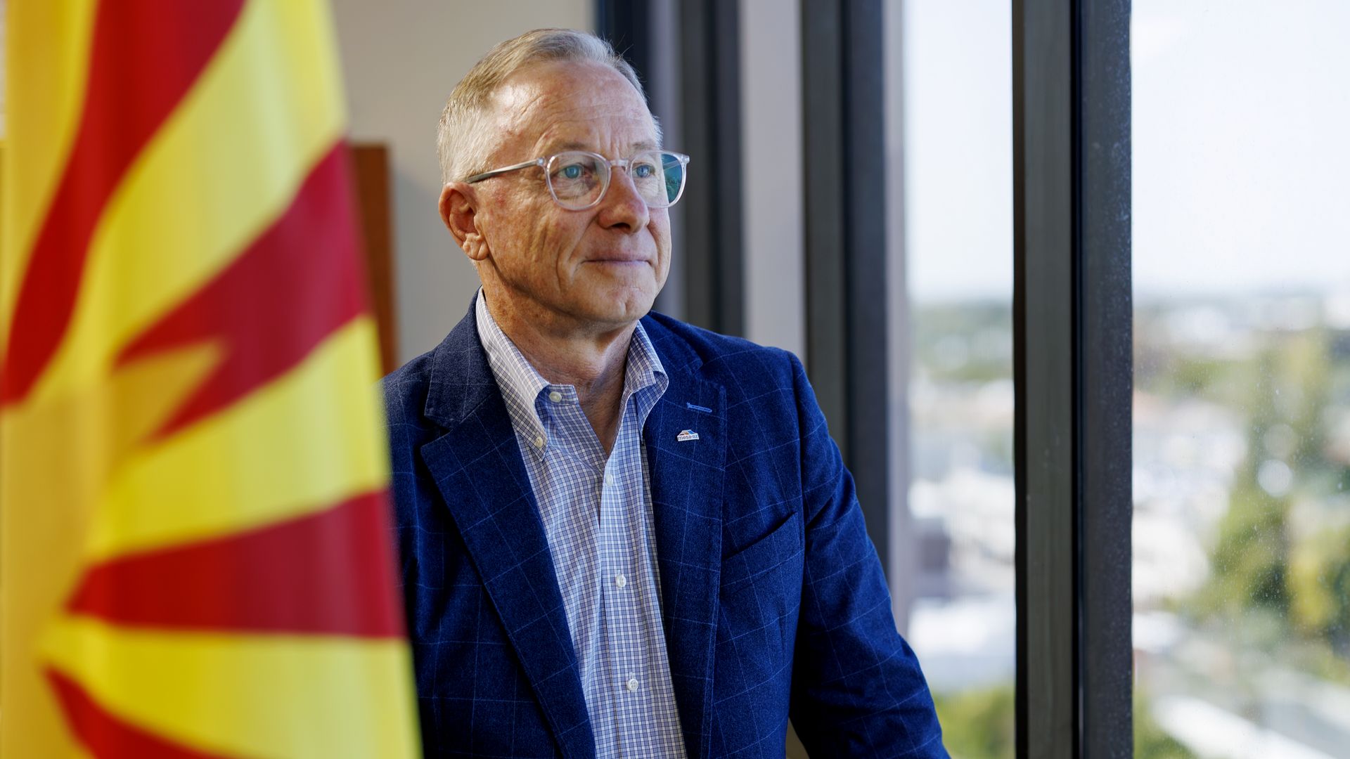 John Giles looks out a window wearing a blue suit with no tie. An Arizona flag is in the foreground