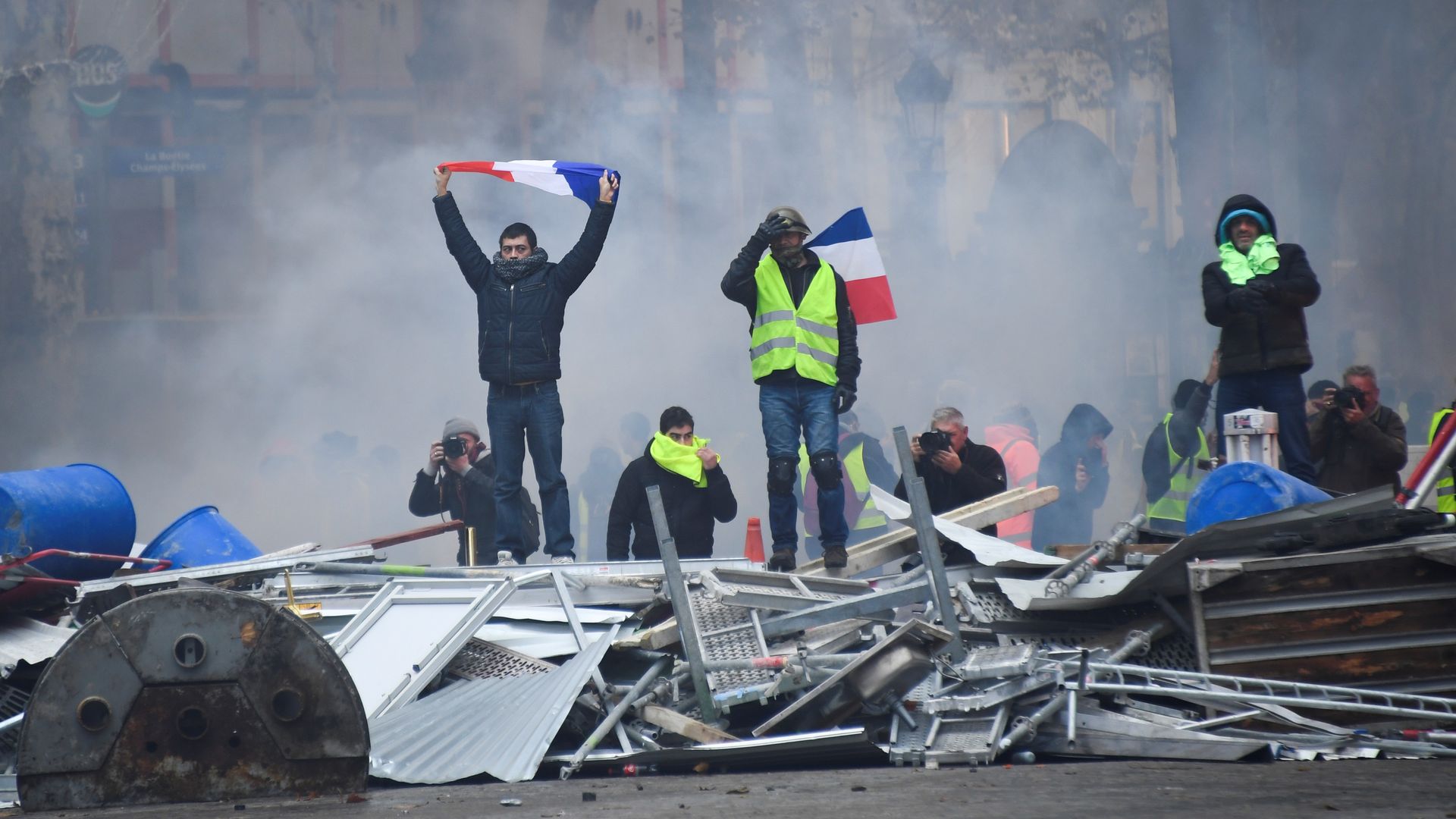 A protestor stands on a pile of debris while holding a French flag over his head.