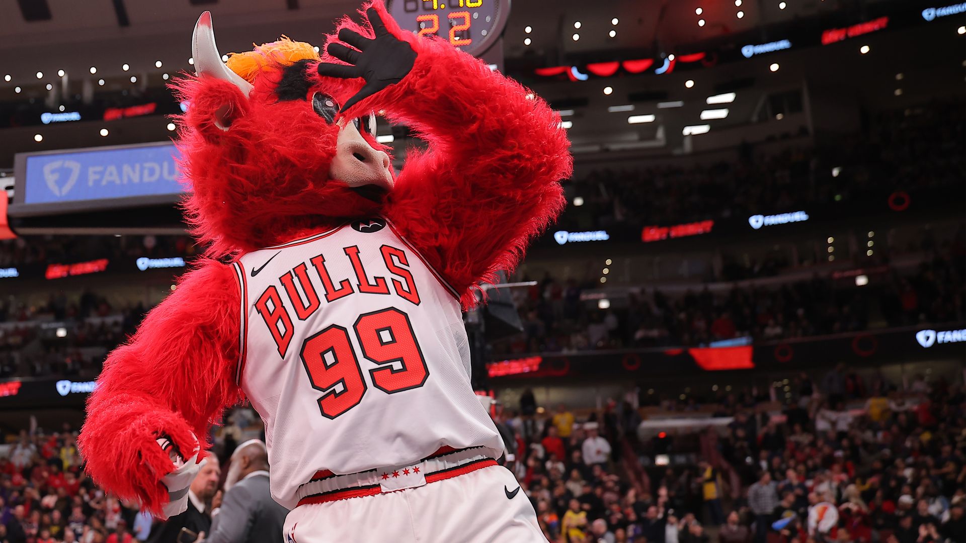 Red furry Chicago Bulls mascot wearing a white jersey with BULLS 99, on a basketball court making a gesture over his eyes, with a cheering crowd and scoreboard in the background.