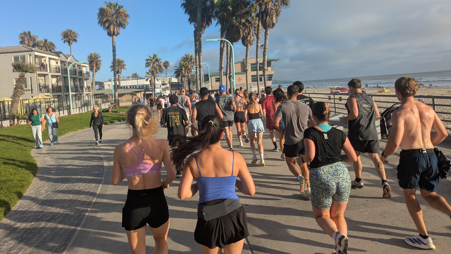 Bright beach boardwalk scene with a large crowd of runners in athletic wear, tall palm trees along the path, blue sky and ocean to the right, and low buildings with spectators on the left.