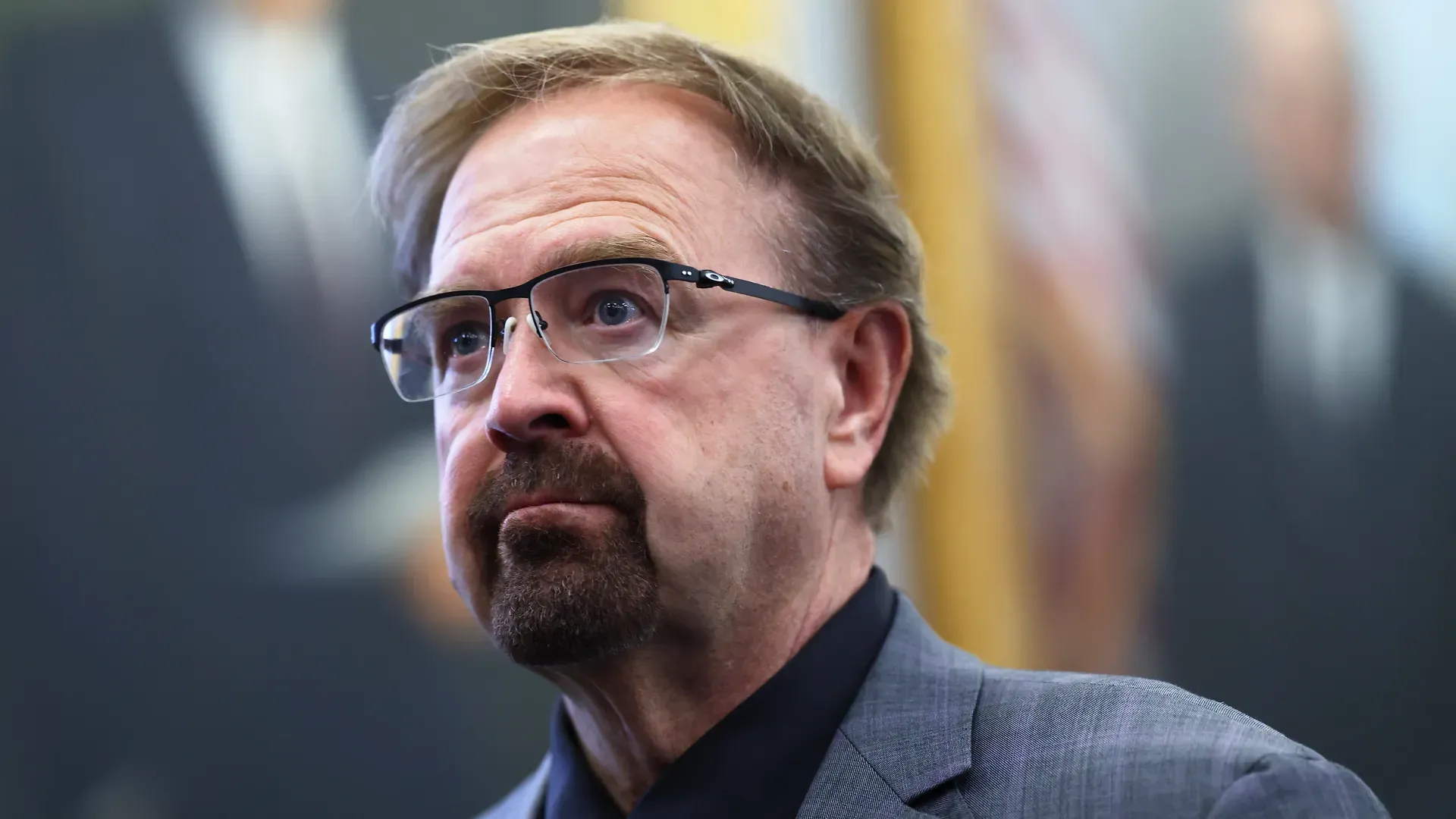 Close-up of a middle-aged man with light brown hair, glasses, and a trimmed beard wearing a gray suit and black shirt, looking to the left with a neutral expression; blurred indoor background.