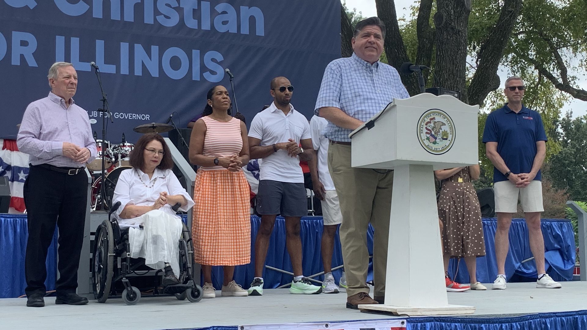 Outdoor political rally: speaker at a podium with the Illinois state seal, a blue backdrop, red‑white‑blue bunting, and diverse supporters including a woman in a wheelchair.