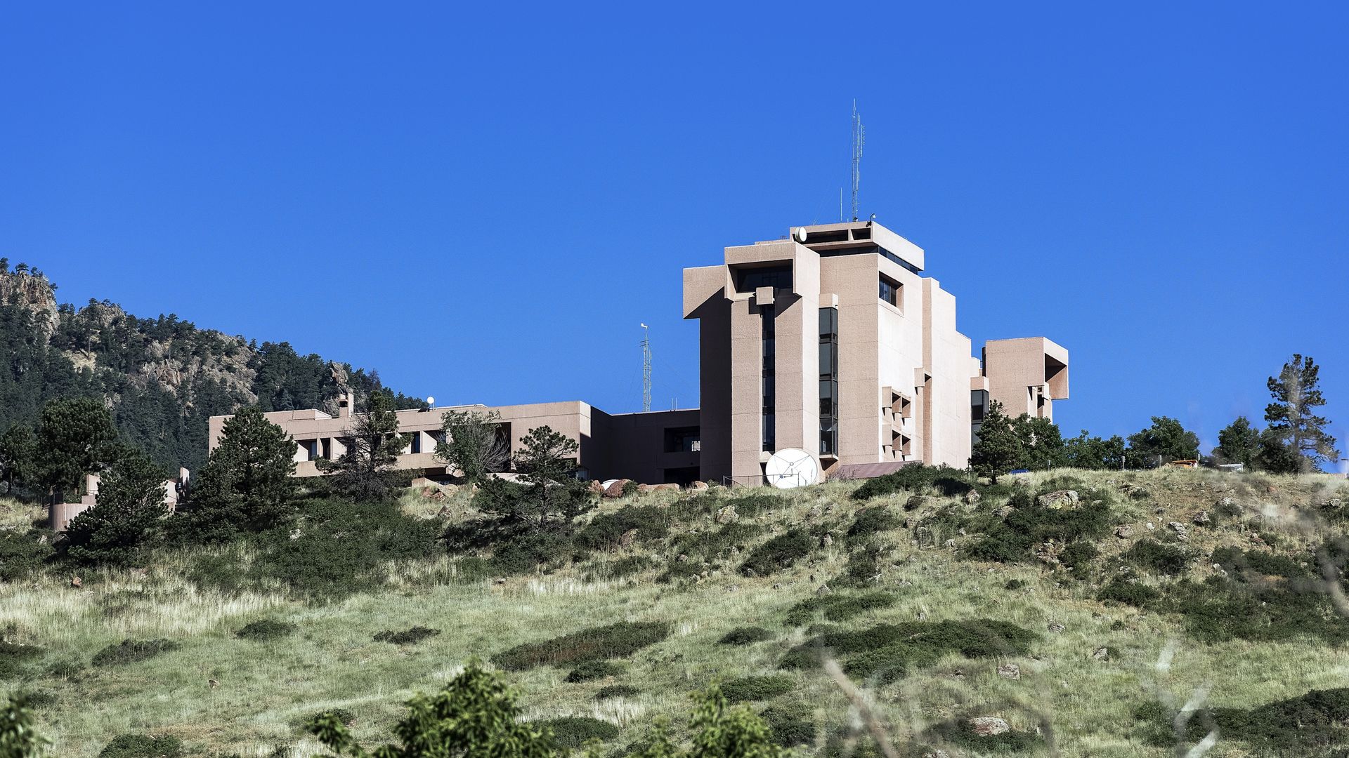Beige multi-story building with satellite dish on grassy hillside under clear blue sky, surrounded by green trees and hill landscape.