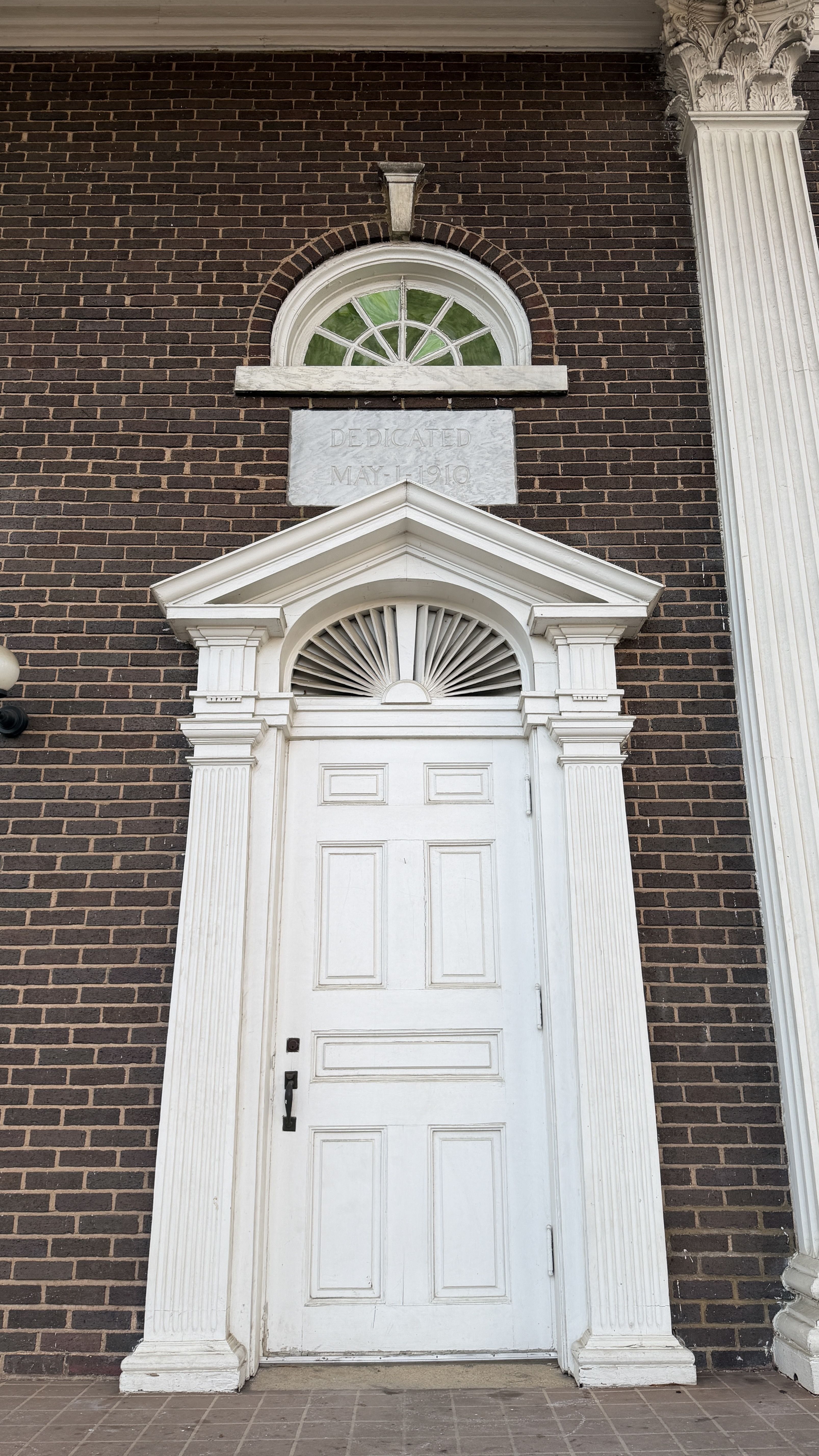 Tall white paneled door with decorative columns and pediment on dark red brick wall, topped by a semicircular window with green glass and marble plaque reading "Dedicated May 1, 1910".
