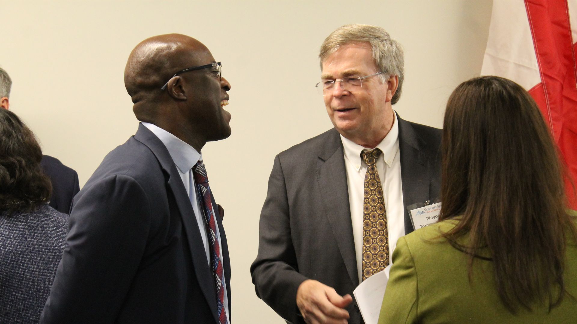 Two suited men converse at a formal event. A bald, dark-skinned man with glasses faces a gray-haired man in a suit and patterned tie; a name badge is visible. Red-and-white flag stands behind them.