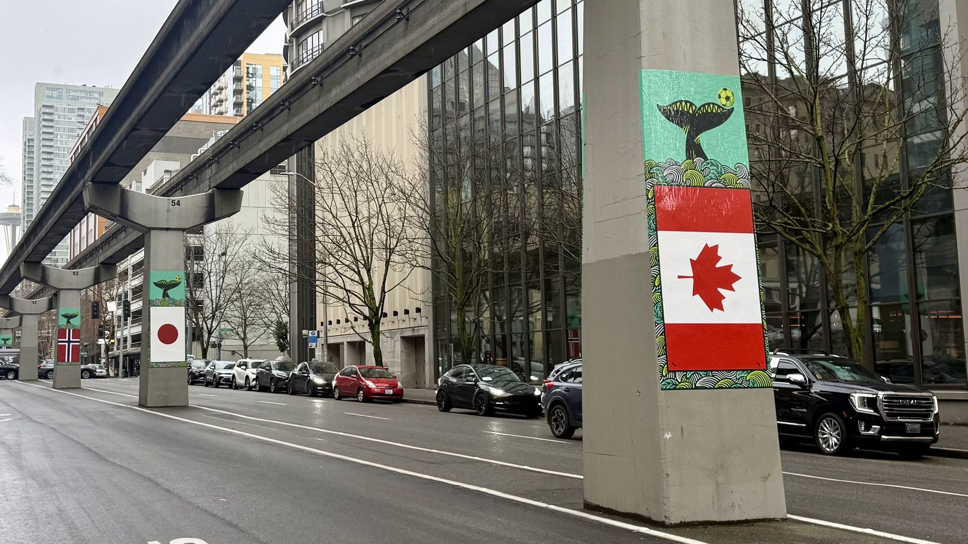 Concrete pillars under a monorail track in a city street, each decorated with flags of Canada, Japan, and Norway, with whale tail and wave artwork on top.