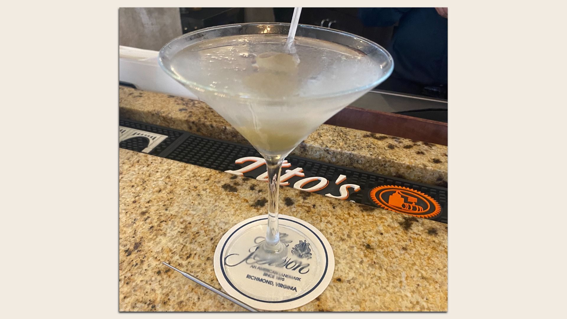 Clear martini glass with a pale cocktail and ice, on a granite bar. A straw sticks up; a round coaster reads Richmond, Virginia, while a black bar mat with an orange logo is in the background.