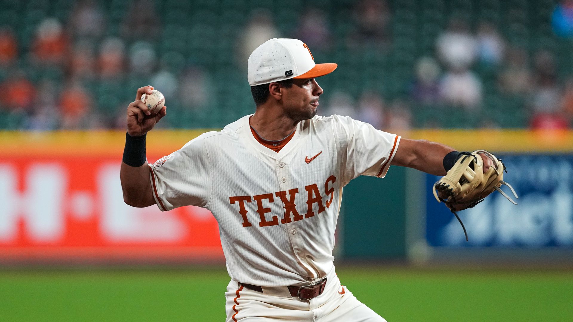 Texas baseball pitcher in a white uniform with orange TEXAS lettering and cap, throwing a pitch with the ball in his left hand and glove on his right, on a green field with blurred stands.