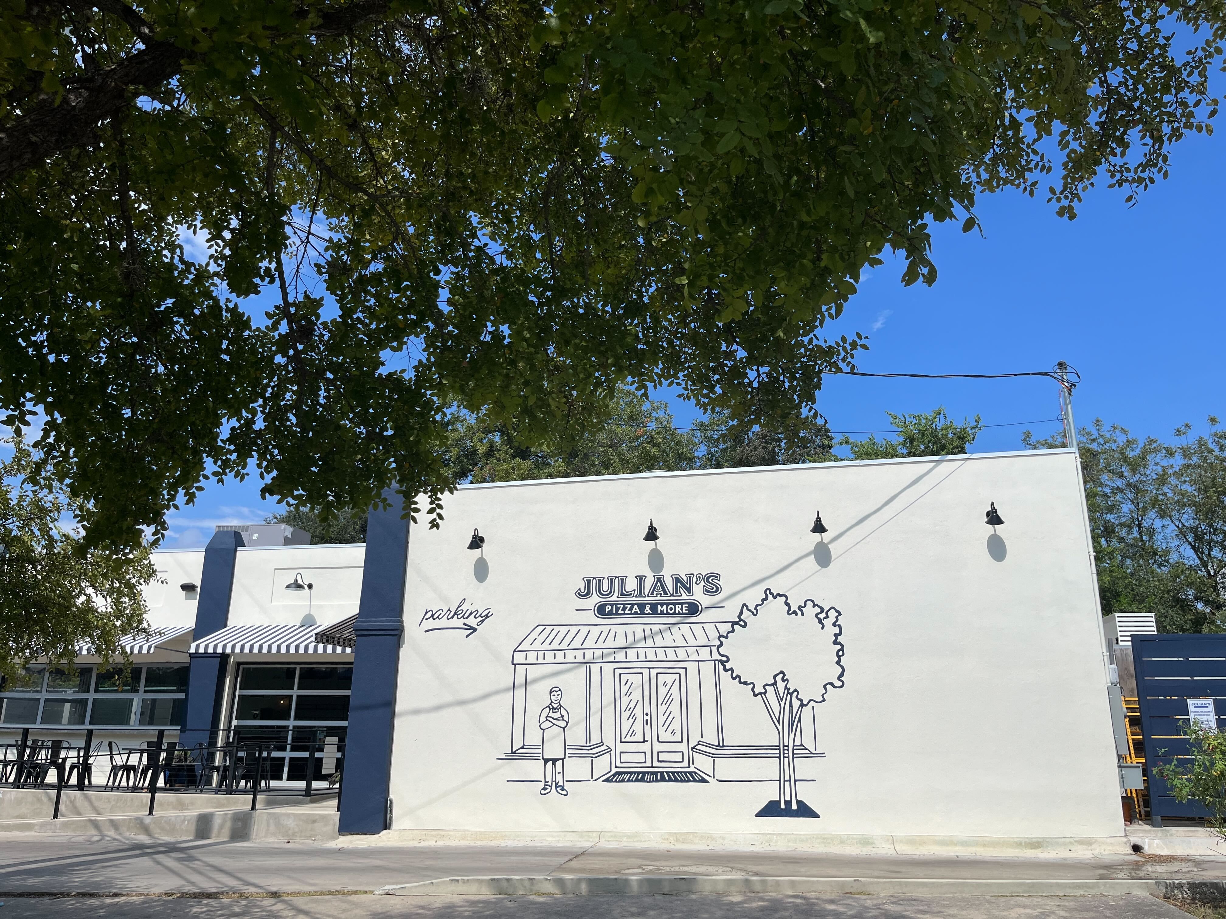 White building with navy accents housing Julian's Pizza & More, featuring a mural of a person, a tree, and parking arrow. Green tree branches frame the blue sky above.
