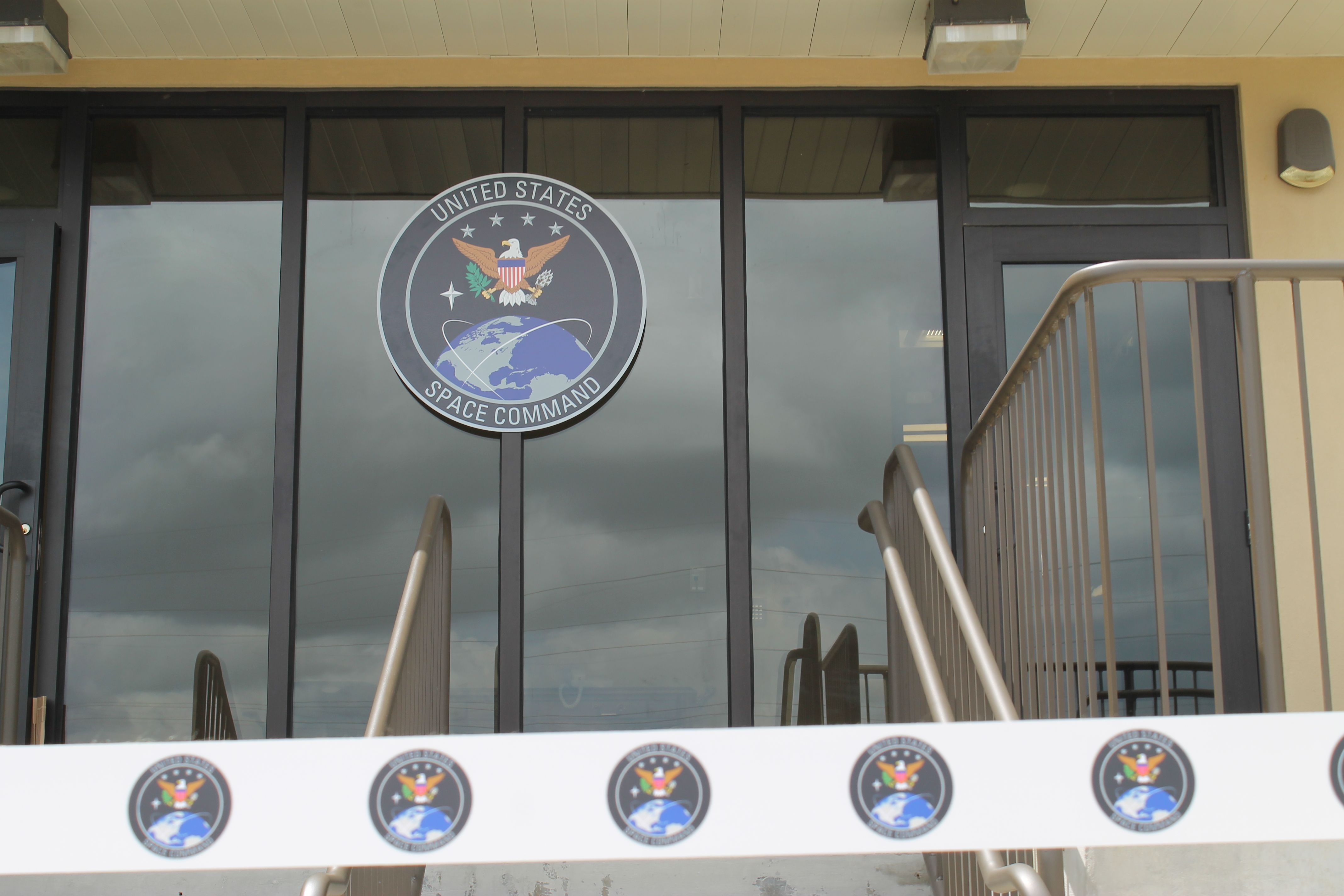 Glass entrance with a large circular United States Space Command emblem on the door, showing an eagle, shield, and globe. A metal staircase railing on the right and cloudy sky reflected in the glass.