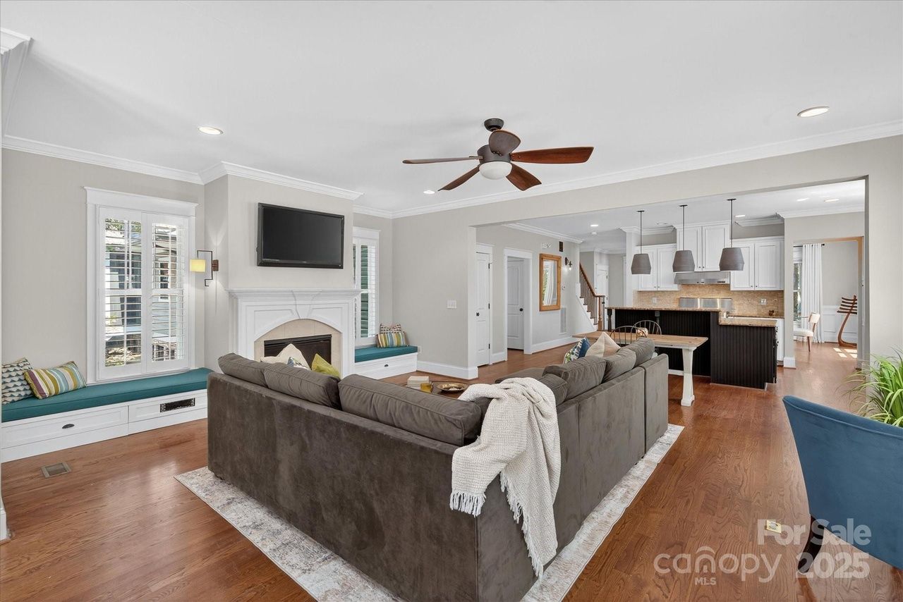 Open living room with a large dark gray sectional sofa, white fireplace with TV above, two window seats with teal cushions, hardwood floors, ceiling fan, and view into white kitchen with dark island.